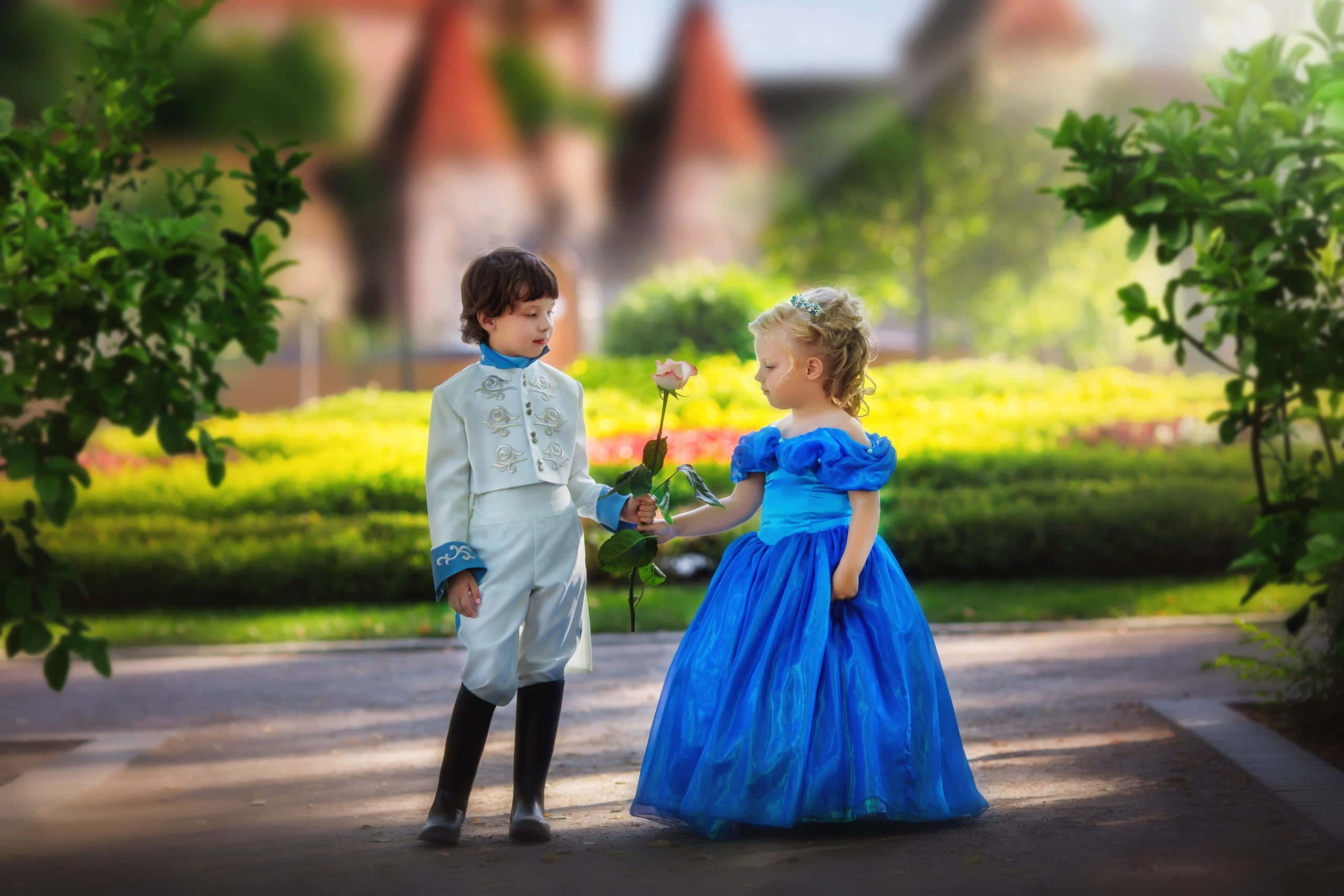 Cute Toddlers 桌布 壁紙 — 一個穿著白色王子服裝的小男孩在陽光明媚的花園裡向一個穿著藍色公主裙的小女孩獻上一朵玫瑰。