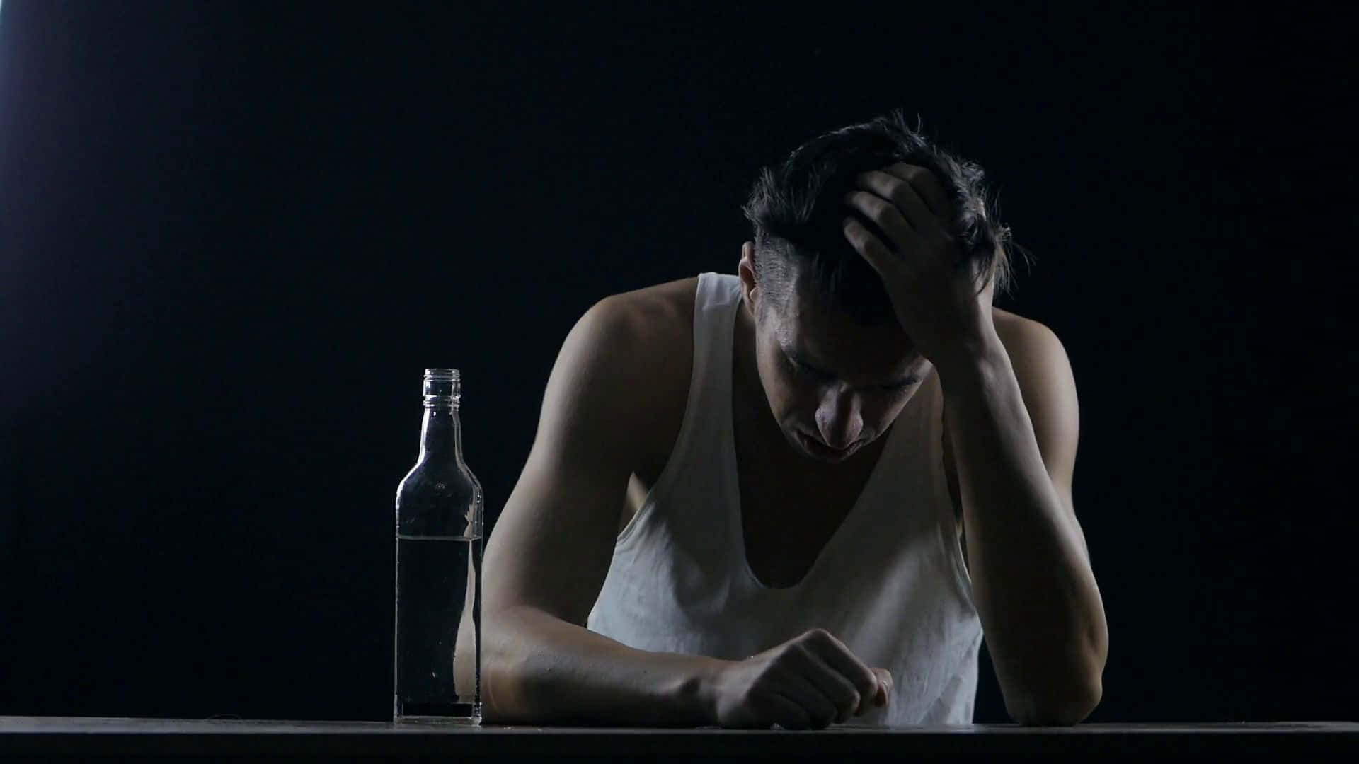 Alone 桌布 壁紙 — 一個穿著白色背心的男人癱坐在一張放著空瓶的桌子旁，雙手抱頭，房間燈光昏暗。