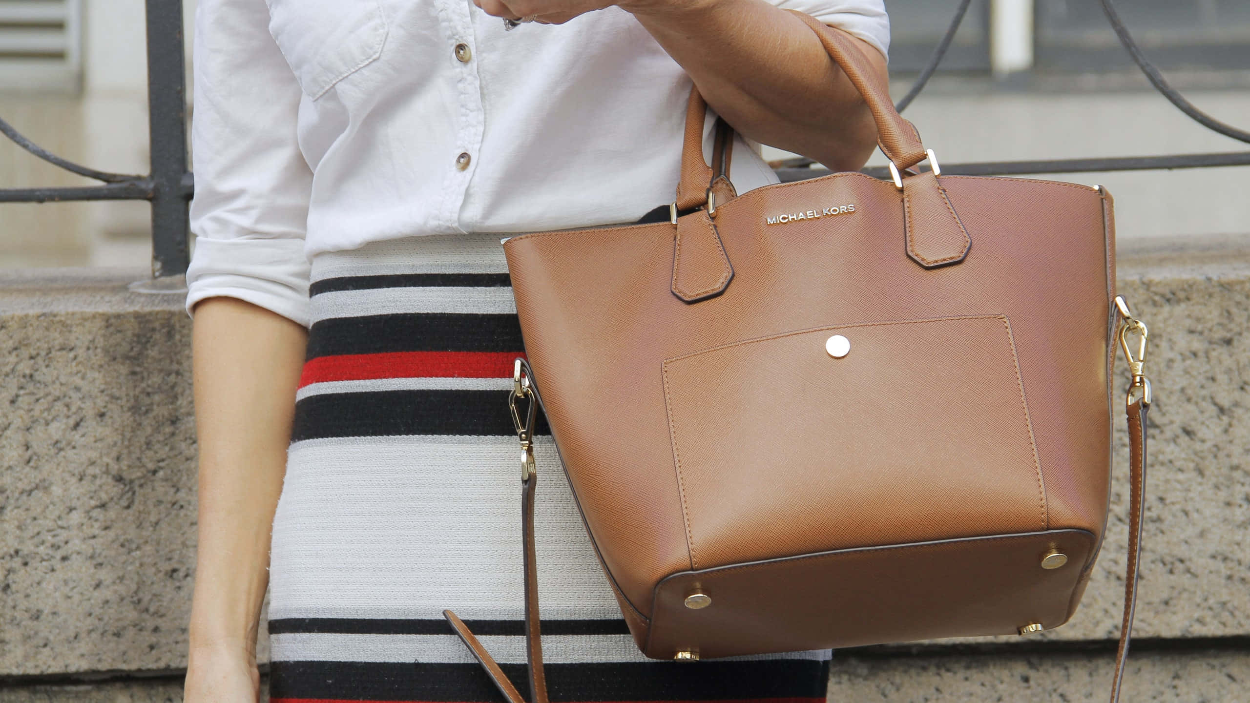 Luxury Brands 桌布 壁紙 — 一個穿著白色襯衫和條紋裙的人，拿著一個棕色皮革Michael Kors手提袋，靠著石牆。