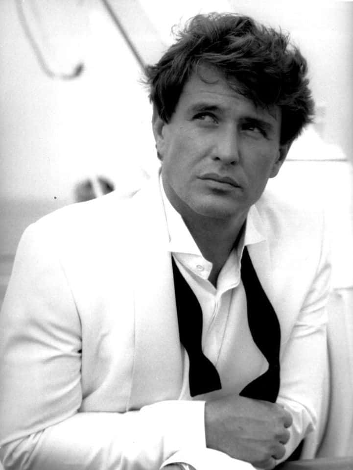 Tom Berenger 桌布 壁紙 — 一個穿著白色西裝的男人，頭髮蓬鬆，若有所思地向上看的黑白肖像。