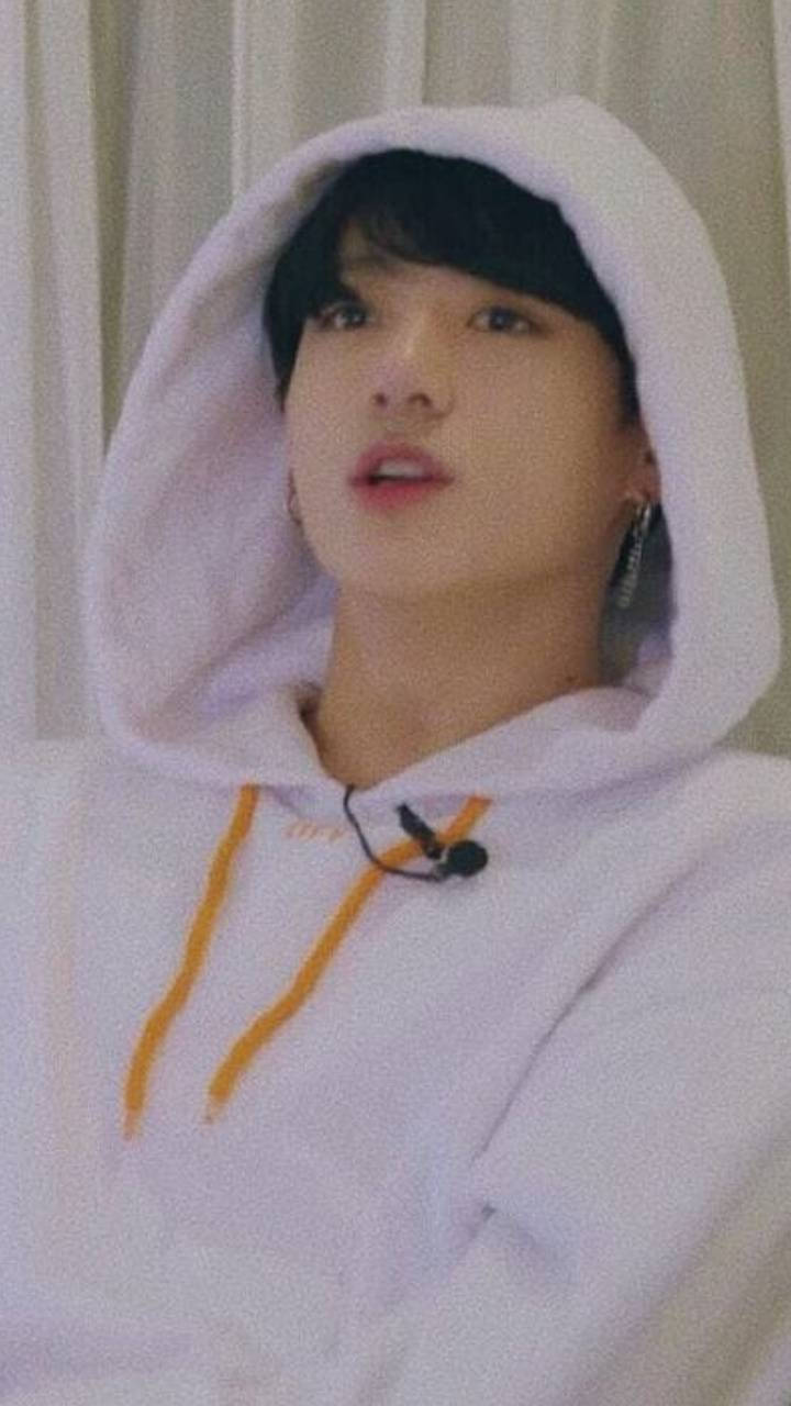 Jungkook Aesthetic 桌布 壁紙 — 一個穿著白色連帽衫、帶橙色抽繩的年輕人，在柔軟的窗簾前抬頭仰望，表情若有所思。