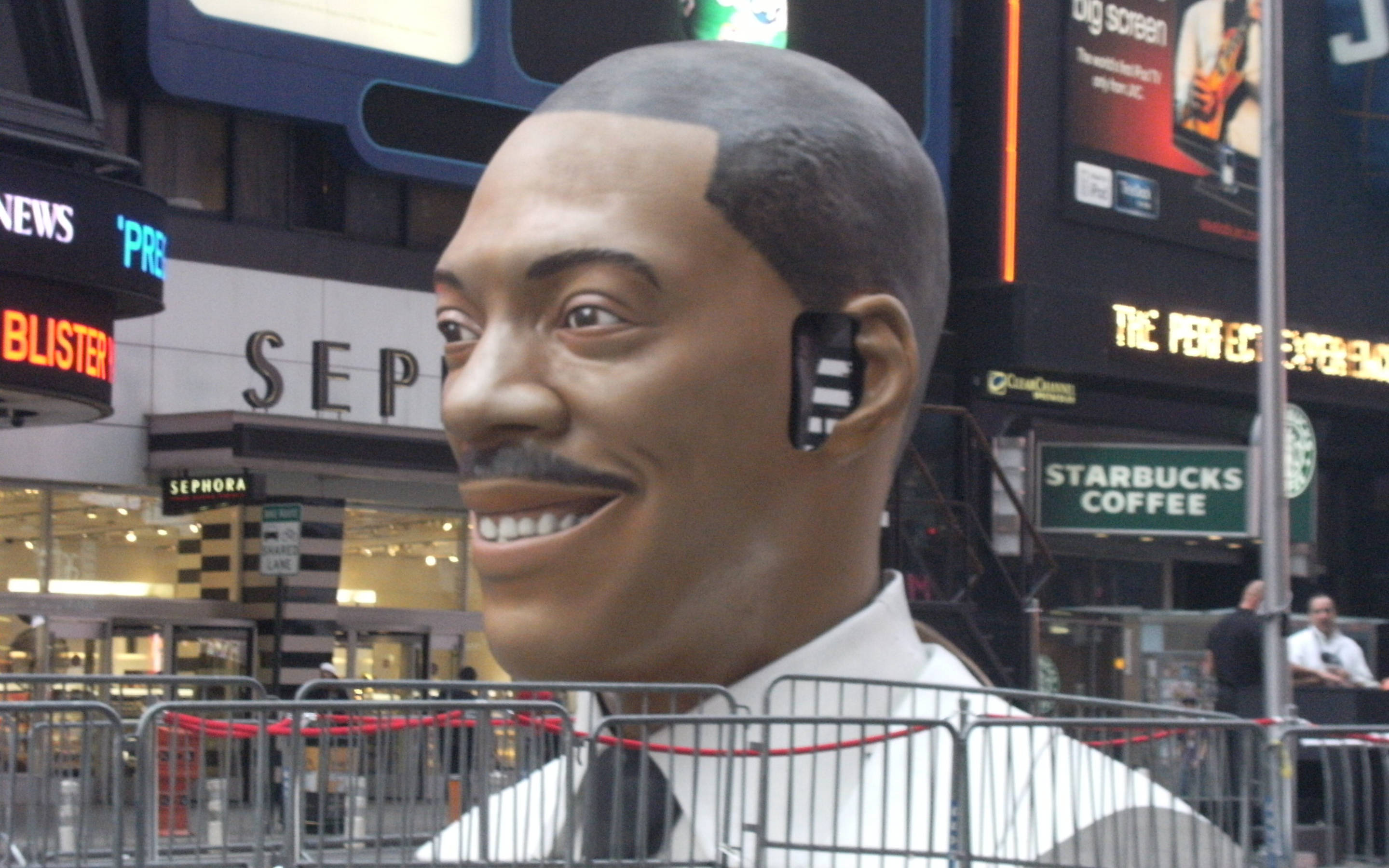 搞笑 桌布 壁紙 — 一個穿著白襯衫打領帶的巨型微笑男子雕像，矗立在繁華的城市街道上，周圍是明亮的廣告牌和金屬護欄。