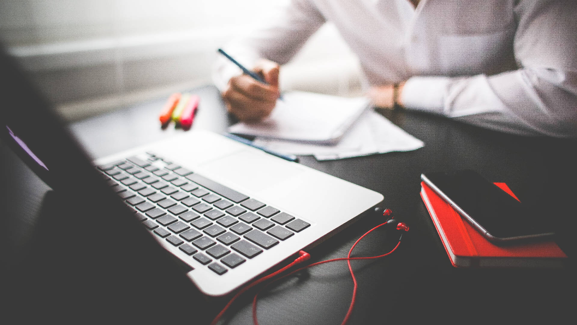 Working 桌布 壁紙 — 一個穿著白襯衫的人正在桌邊寫筆記，旁邊有筆記型電腦、紅色耳機和彩色筆。
