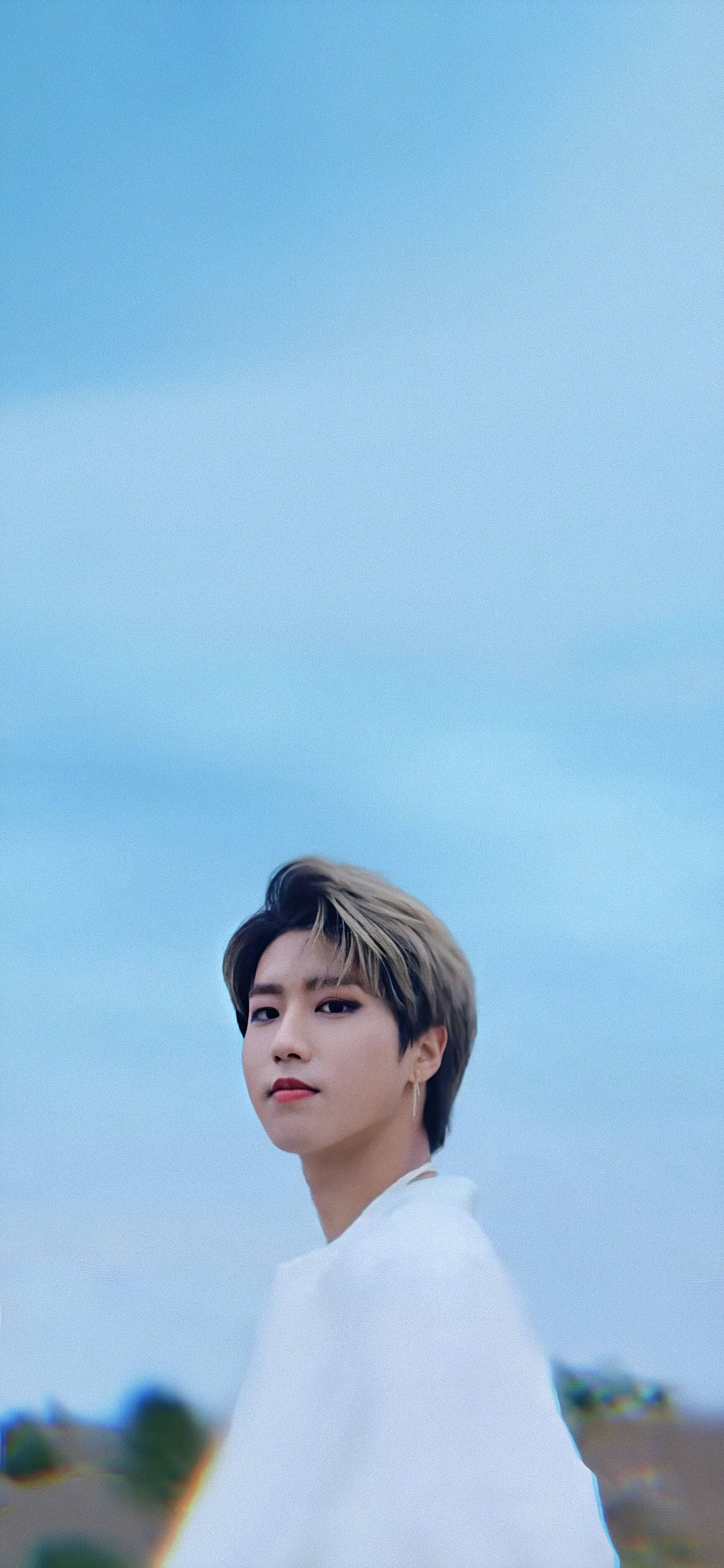 Stray Kids 桌布 壁紙 — 一個穿著白襯衫的年輕男子在柔和的藍天和模糊的遠山背景下回頭凝視。