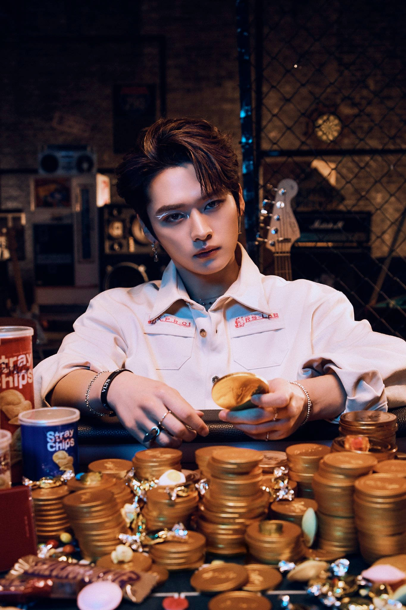 Stray Kids 桌布 壁紙 — 一個穿著白襯衫的年輕男子坐在一張堆滿金幣和零食罐的桌子旁，在一個光線昏暗的車庫式房間裡。