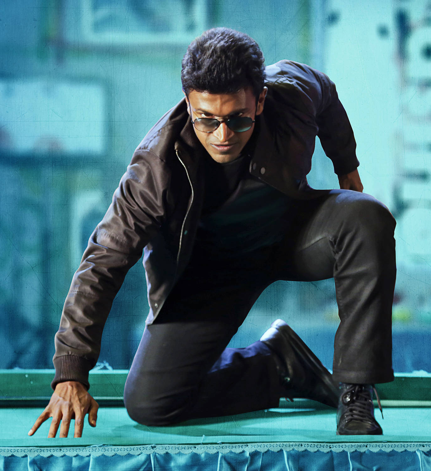 Puneeth Rajkumar 桌布 壁紙 — 一個穿著皮夾克和太陽眼鏡的男人在青綠色表面上低蹲，背景是粗糙的城市景觀。