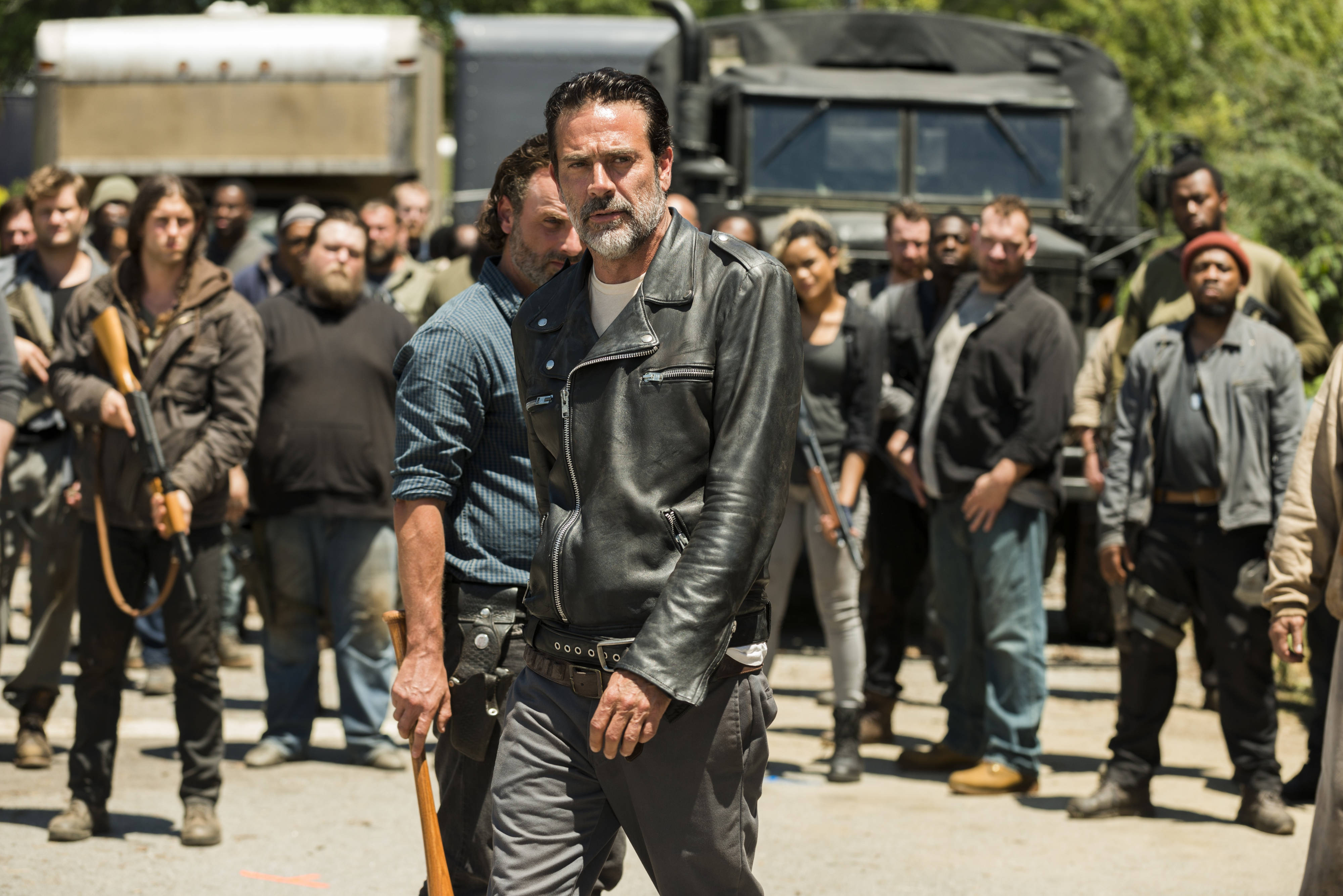 Negan 桌布 壁紙 — 一個穿著皮夾克的嚴肅男人站在畫面中央，手持球棒，而一群武裝倖存者在明亮的日光下隱約出現在他身後。