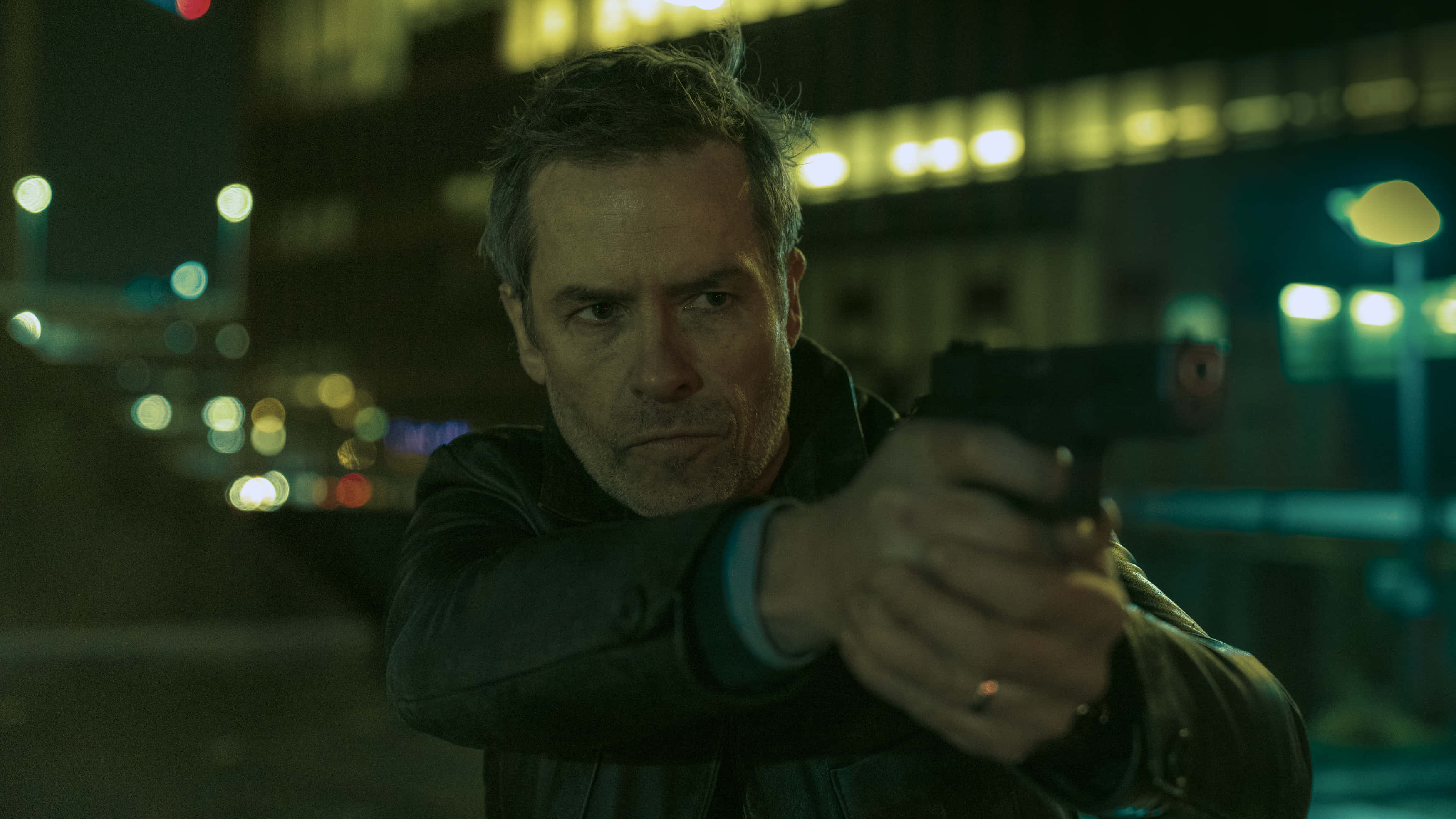Guy Pearce 桌布 壁紙 — 一個穿著皮夾克的男人，在模糊的霓虹燈城市夜景背景下，專注地瞄準一把手槍。