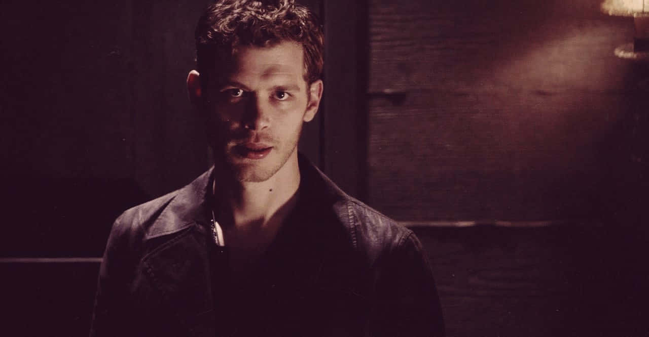 Vampire Diaries 桌布 壁紙 — 一個穿著皮夾克的男人在溫暖的聚光燈下，對著深色木牆專注地凝視。