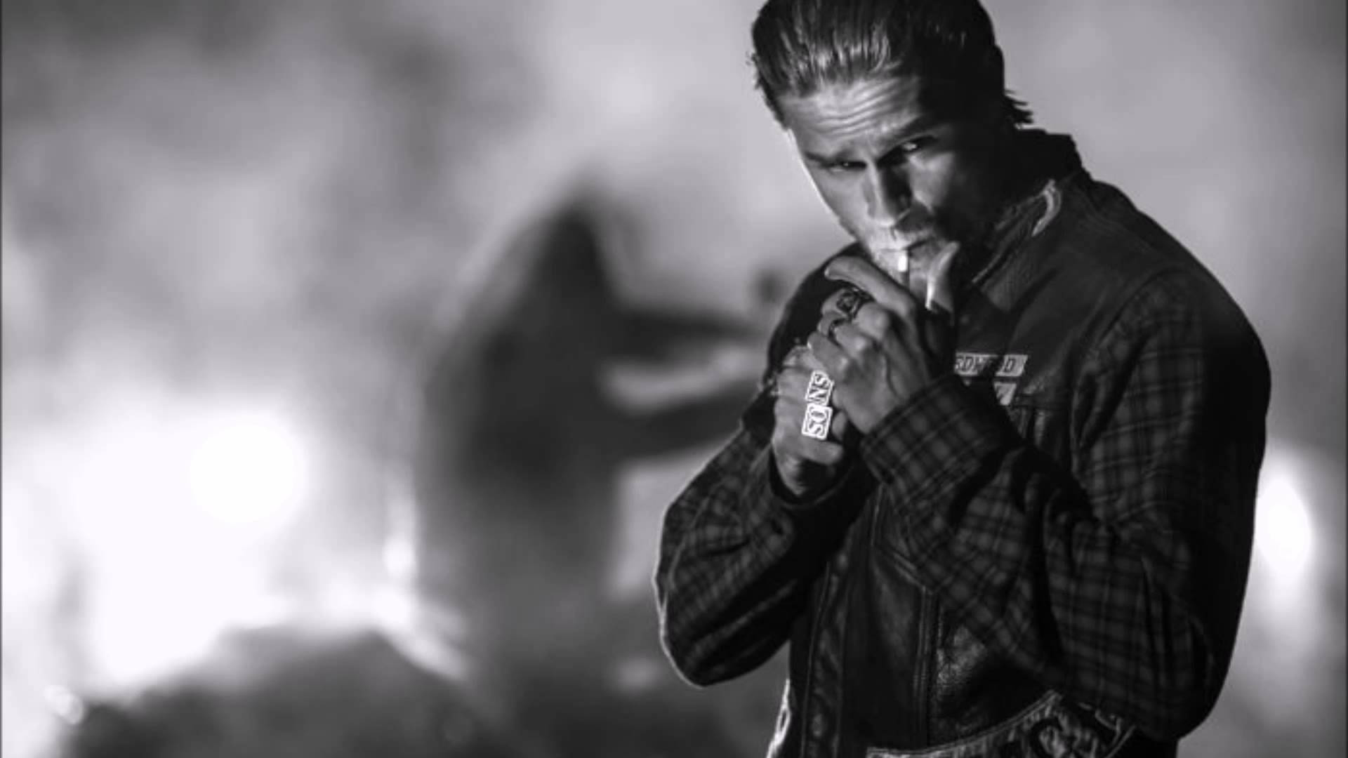 Sons Of Anarchy 桌布 壁紙 — 一個穿著皮夾克的男人在煙霧瀰漫的背景中抽煙，背景有模糊的人影和明亮的燈光。