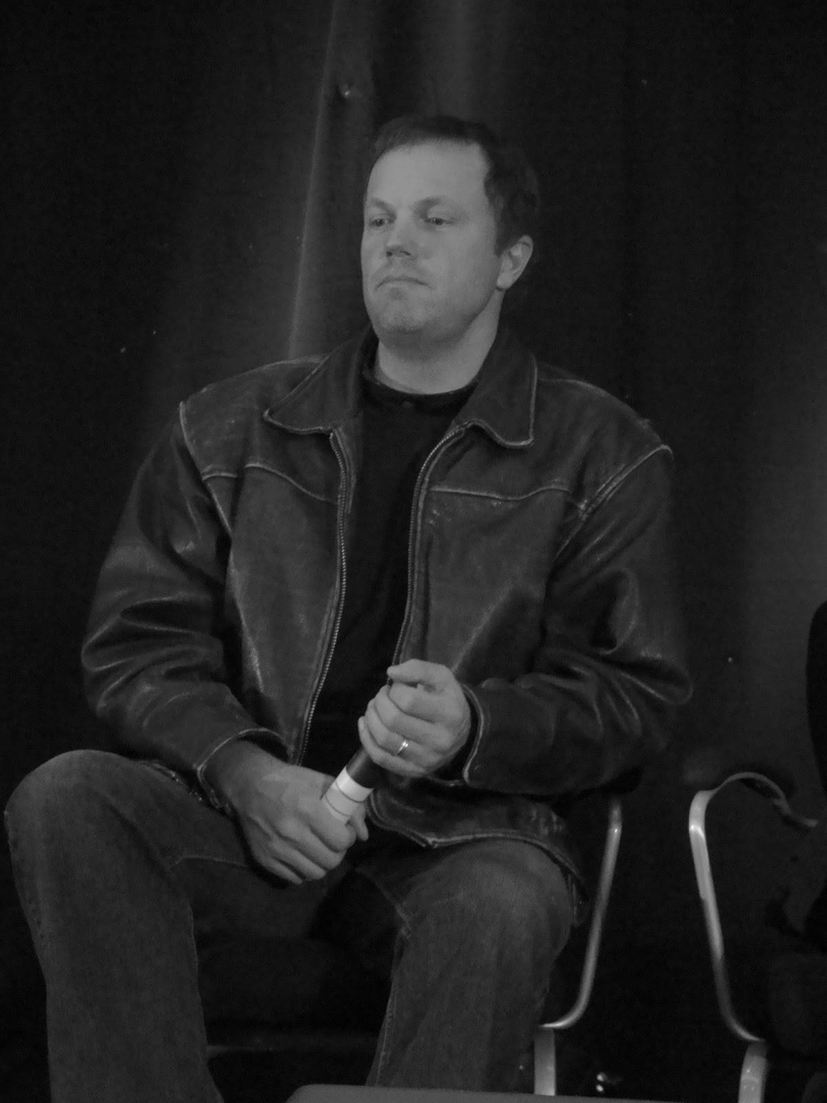 adam baldwin 桌布 壁紙 — 一個穿著皮夾克的男人坐在椅子上，對著黑色窗簾沉思，手持麥克風。