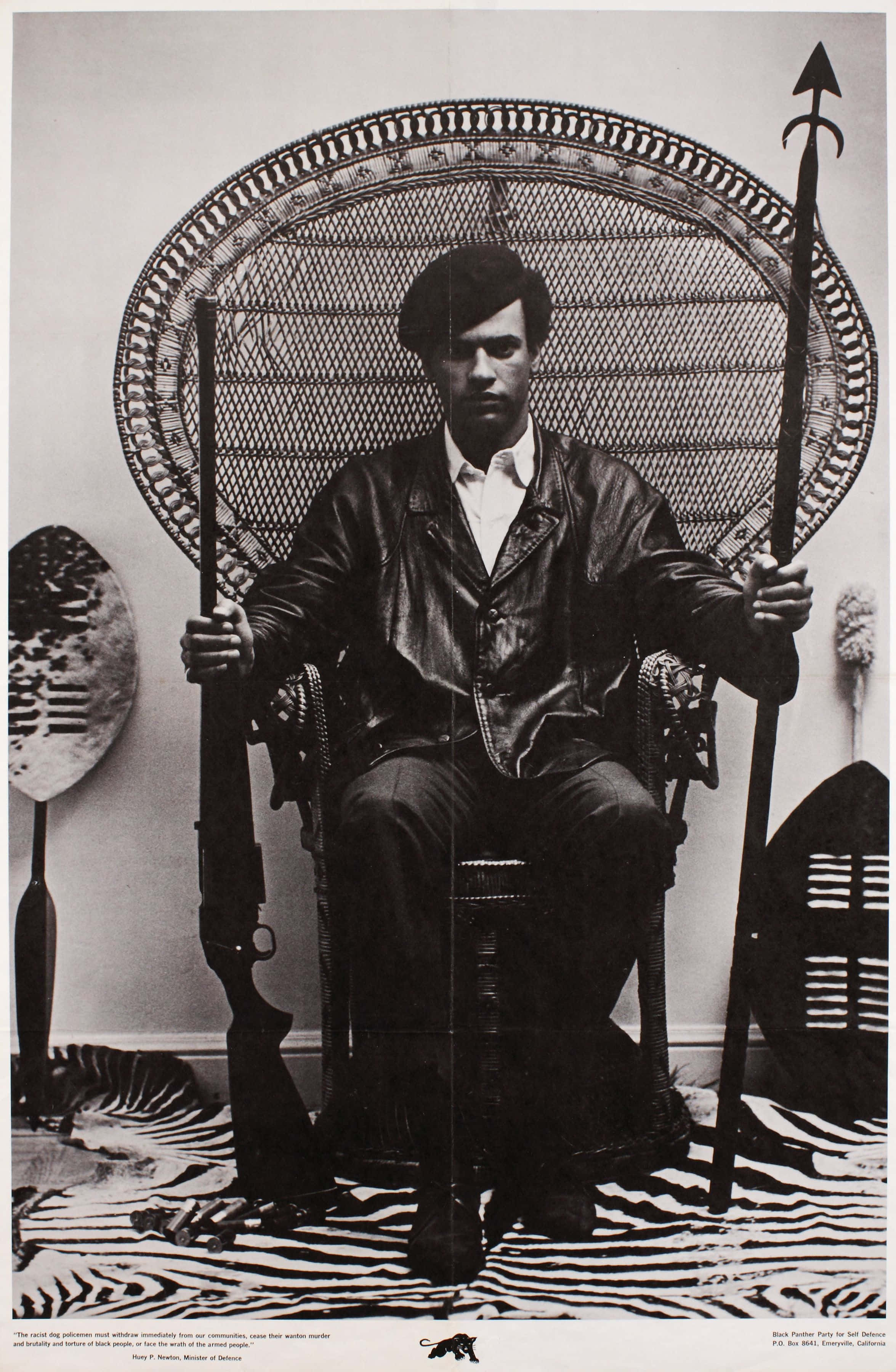 Black Panther Party 桌布 壁紙 — 一個穿著皮夾克的男人威嚴地坐在一張大型編織椅上，手持長矛，兩側有部落盾牌，腳下是斑馬紋地毯。