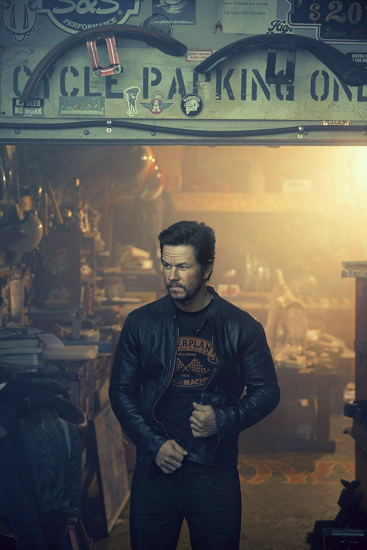 Mark Wahlberg 桌布 壁紙 — 一個穿著皮夾克的男人站在一個光線昏暗的車庫裡，車庫裡充滿了機車零件和復古標誌。