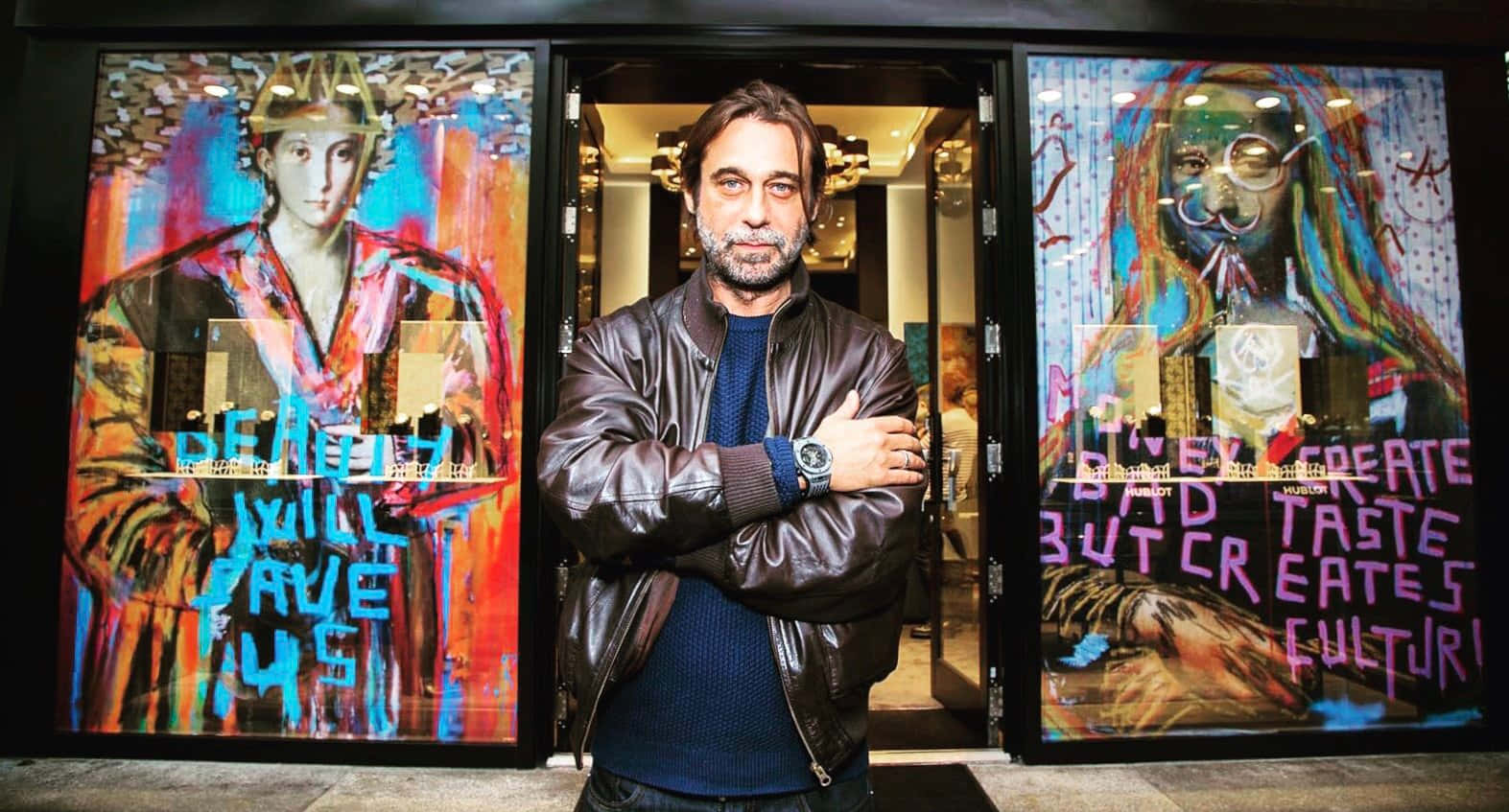 Jordi Mollà 桌布 壁紙 — 一個穿著皮夾克的男人自信地交叉雙臂，站在充滿活力的塗鴉玻璃門前。