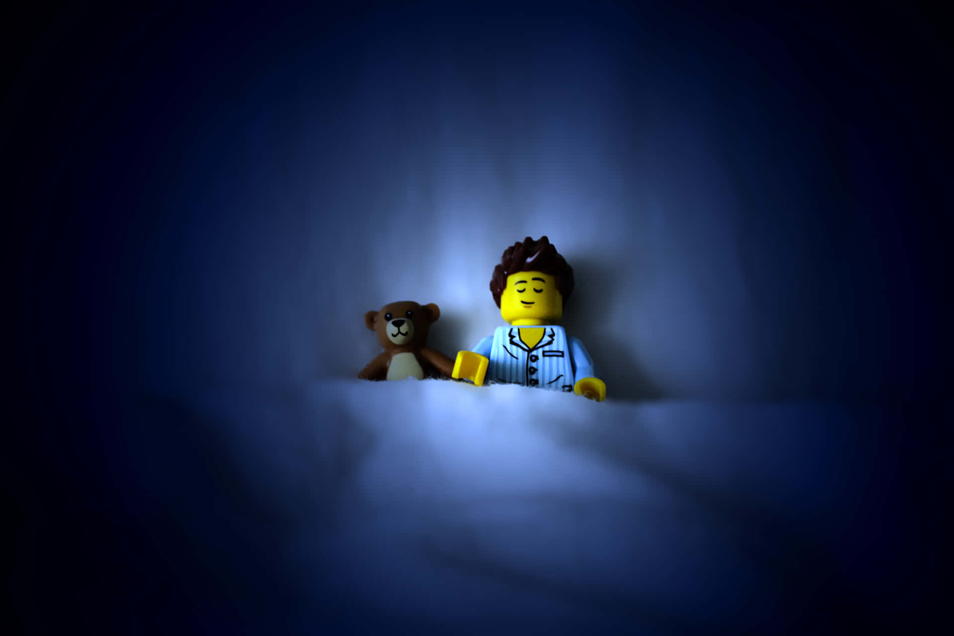 樂高 桌布 壁紙 — 一個穿著睡衣的LEGO迷你人偶在黑暗的房間裡，在柔和的藍光下，在泰迪熊旁邊安詳地睡覺。