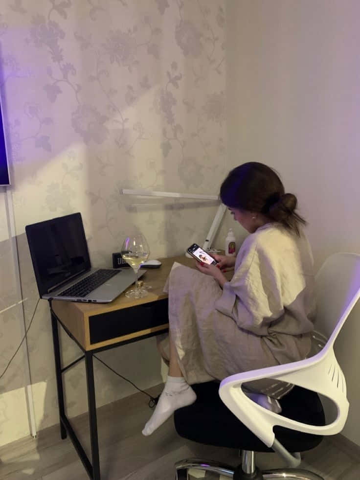 Studying 桌布 壁紙 — 一個穿著睡袍的人坐在書桌前，房間燈光柔和，壁紙是花卉圖案，旁邊有筆記型電腦和酒杯，正在使用電話。