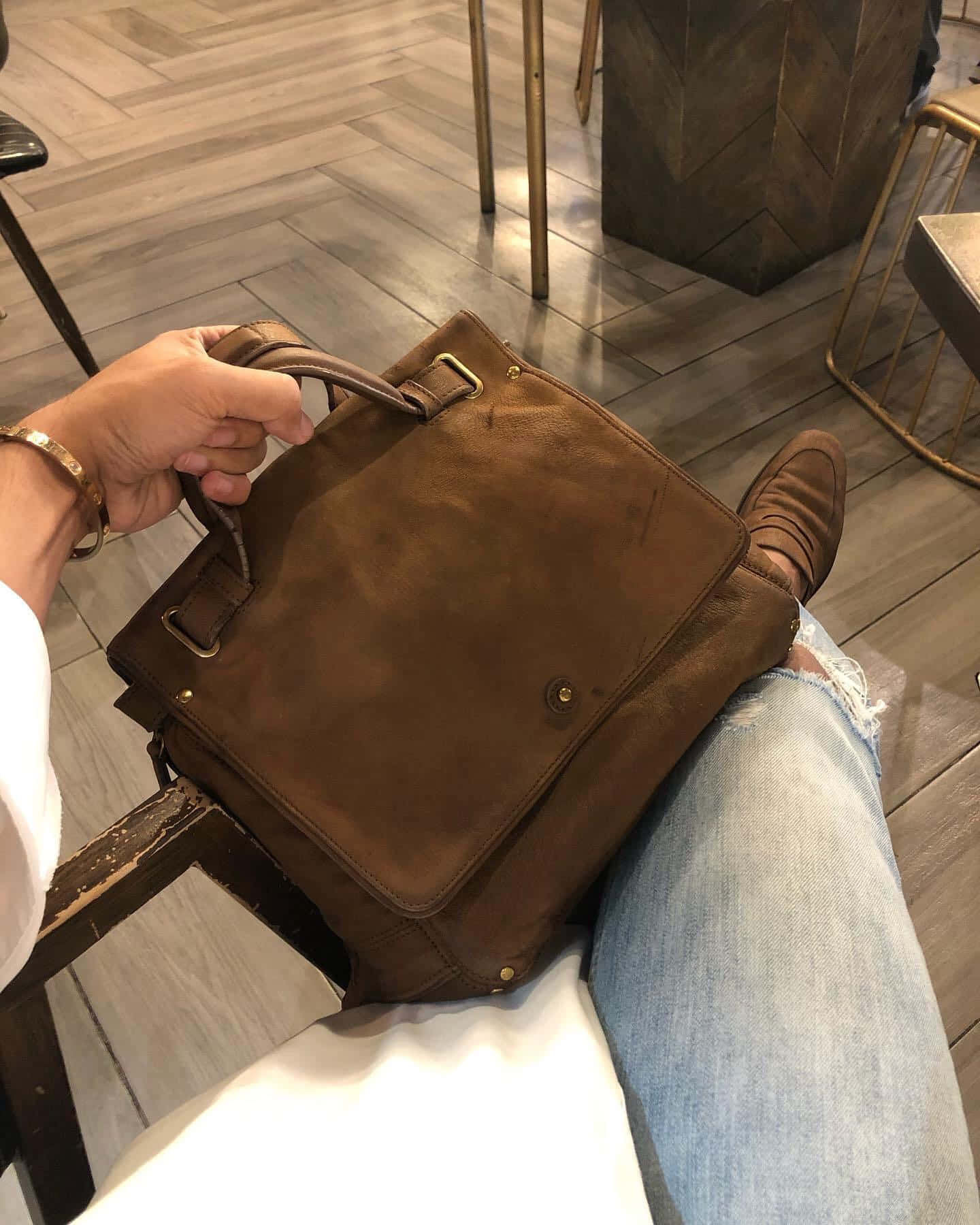 Handbag 桌布 壁紙 — 一個穿著破洞牛仔褲和樂福鞋的人，在咖啡館的木地板上拿著一個棕色麂皮公事包。
