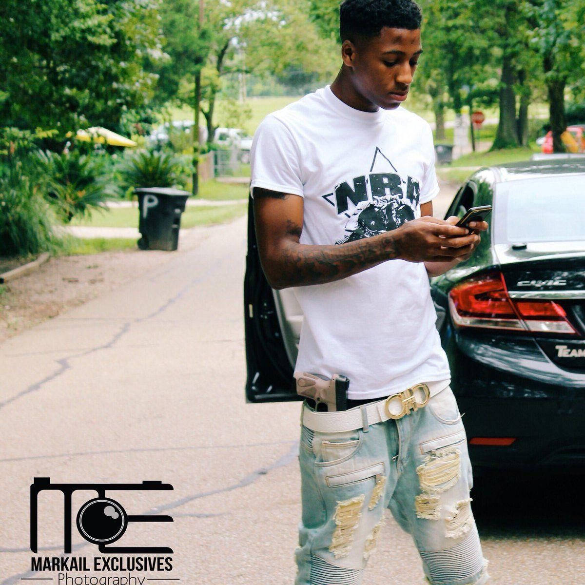 Youngboy 桌布 壁紙 — 一個穿著破洞牛仔褲和白色T恤的年輕男子站在一輛黑色汽車旁，在綠樹成蔭的郊區街道上專注於他的手機。