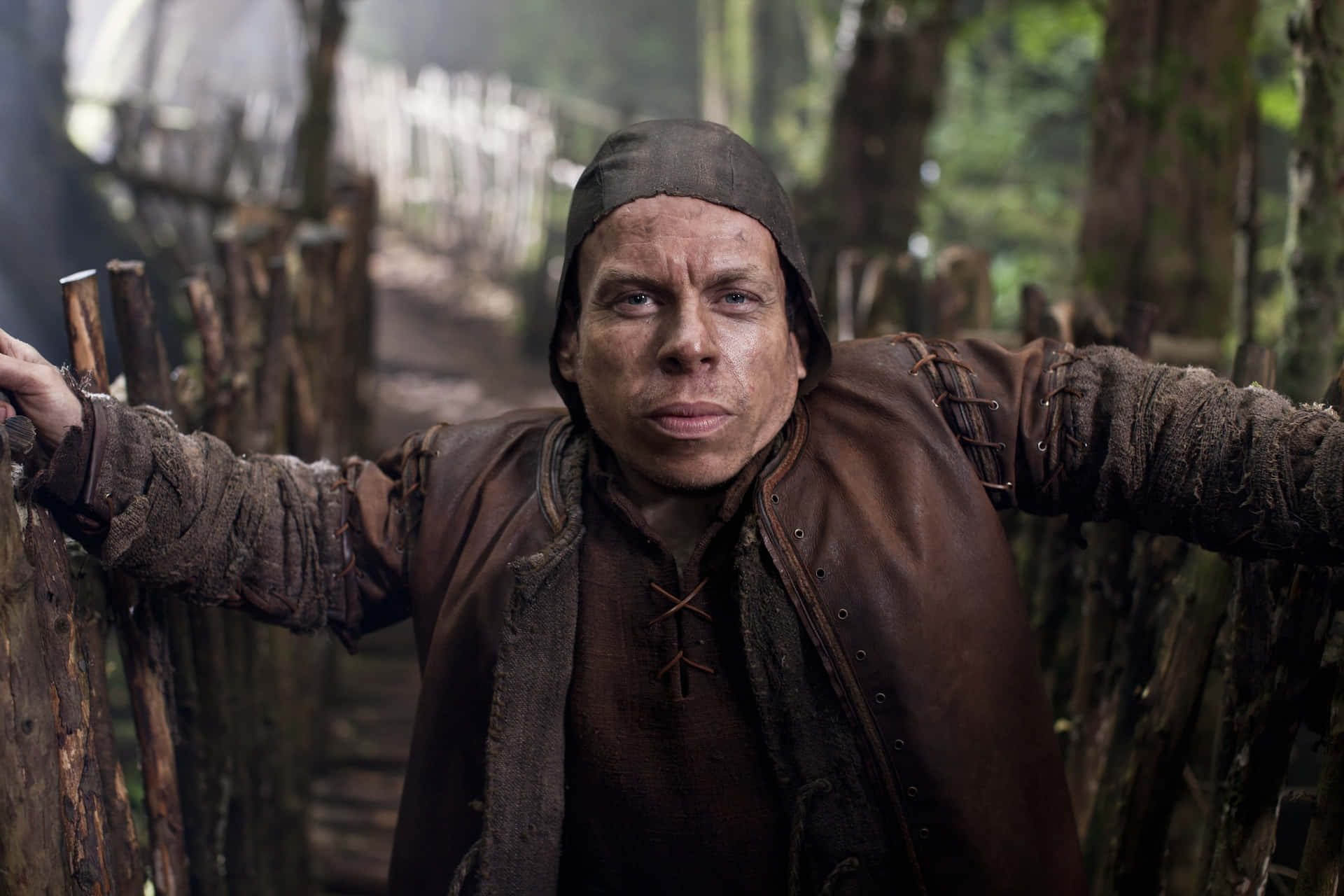 Warwick Davis 桌布 壁紙 — 一個穿著破爛中世紀服裝的男人，在迷霧森林中帶著警覺的表情靠在木柵欄上。
