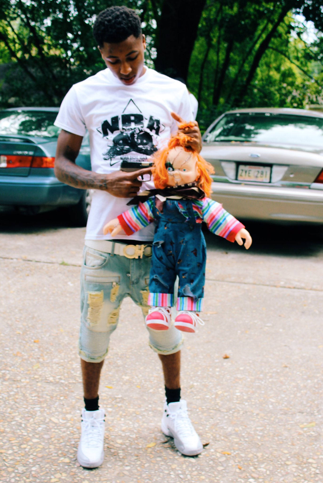 Youngboy 桌布 壁紙 — 一個穿著破爛牛仔褲和白色運動鞋的年輕男子，手持色彩繽紛的Chucky娃娃，站在鋪砌的街道上，身後有停泊的汽車和樹木。