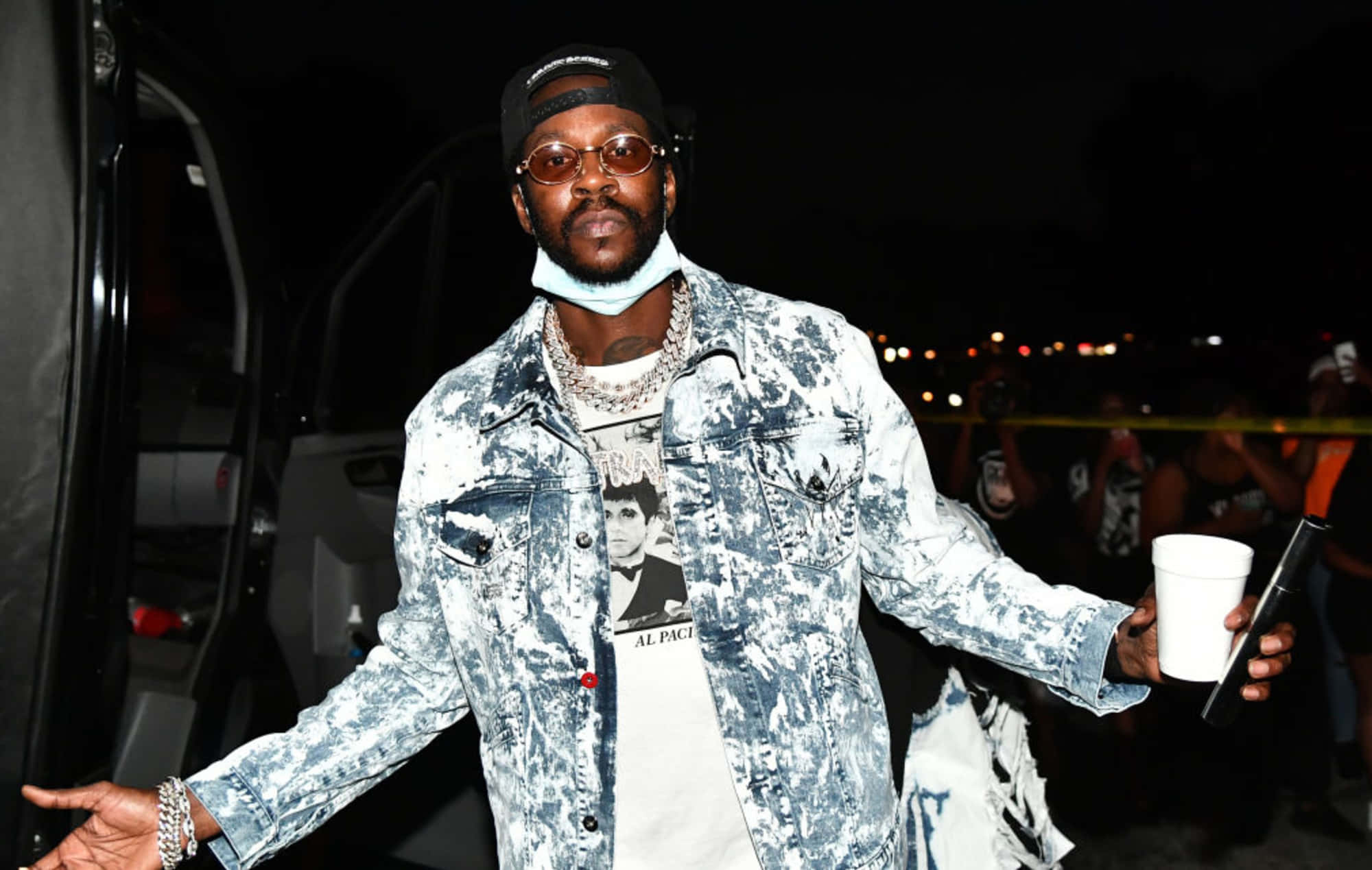 2 Chainz 桌布 壁紙 — 一個穿著破舊牛仔外套和帽子的男人在夜晚自信地站著，手拿杯子和電話，背後的城市燈光閃爍。