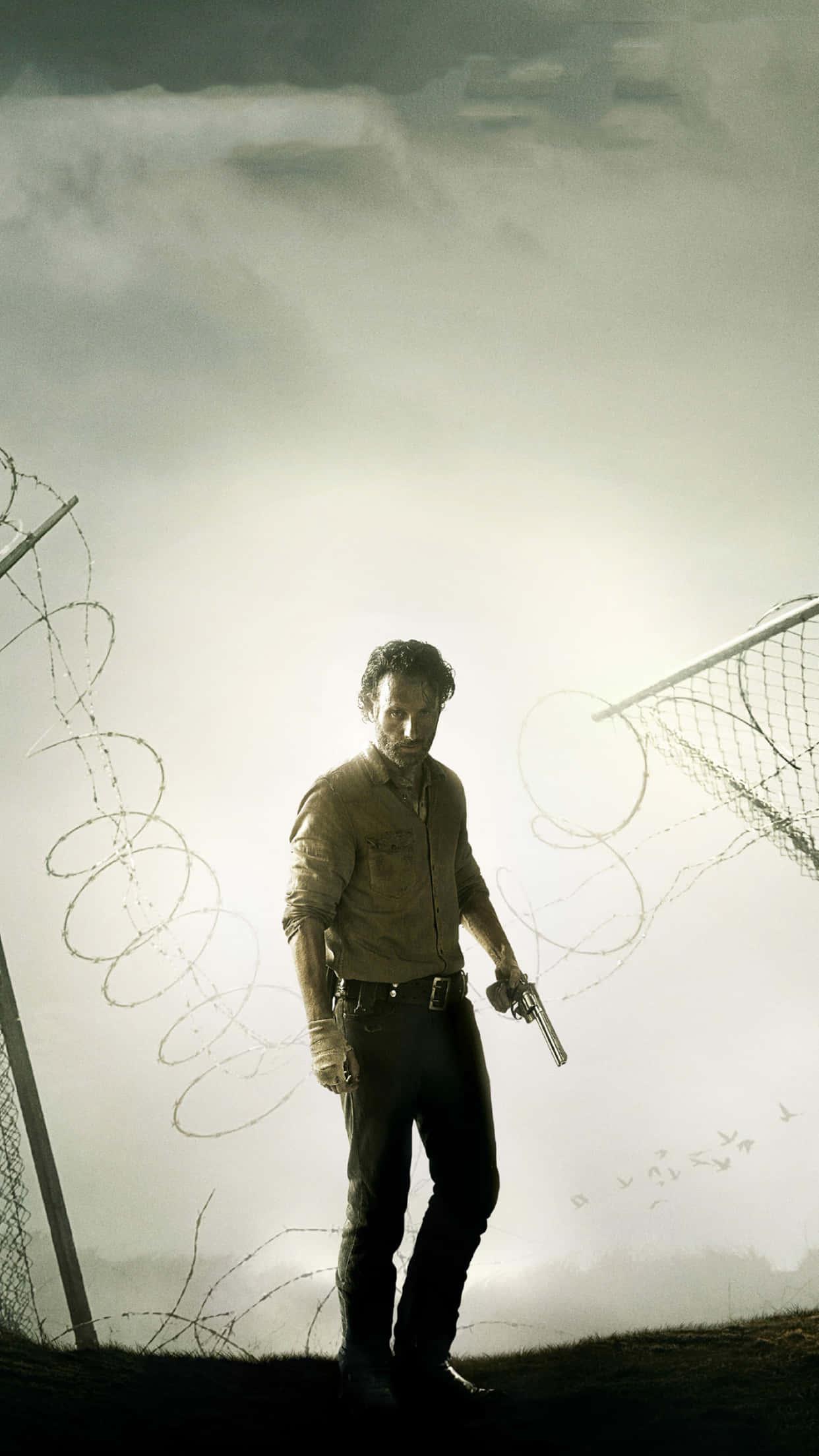 The Walking Dead iPhone 桌布 壁紙 — 一個穿著破舊襯衫的男人，在朦朧的天空下，挑釁地拿著手槍站在破損的鐵絲網圍欄旁。