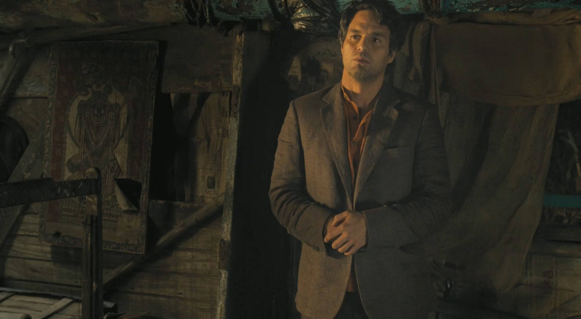 Mark Ruffalo 桌布 壁紙 — 一個穿著破舊西裝的男人站在一個光線昏暗、雜亂的房間裡，房間有木牆和掛著的掛毯，在溫暖的燈光下。