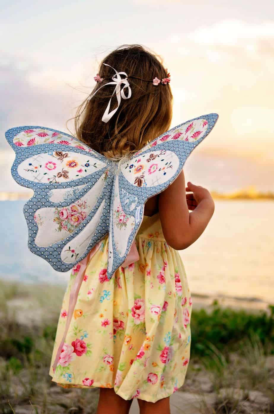 Butterfly Wing Dress 桌布 壁紙 — 一個穿著碎花裙和蝴蝶翅膀的小女孩站在日落時分的沙灘上，凝視著水面。