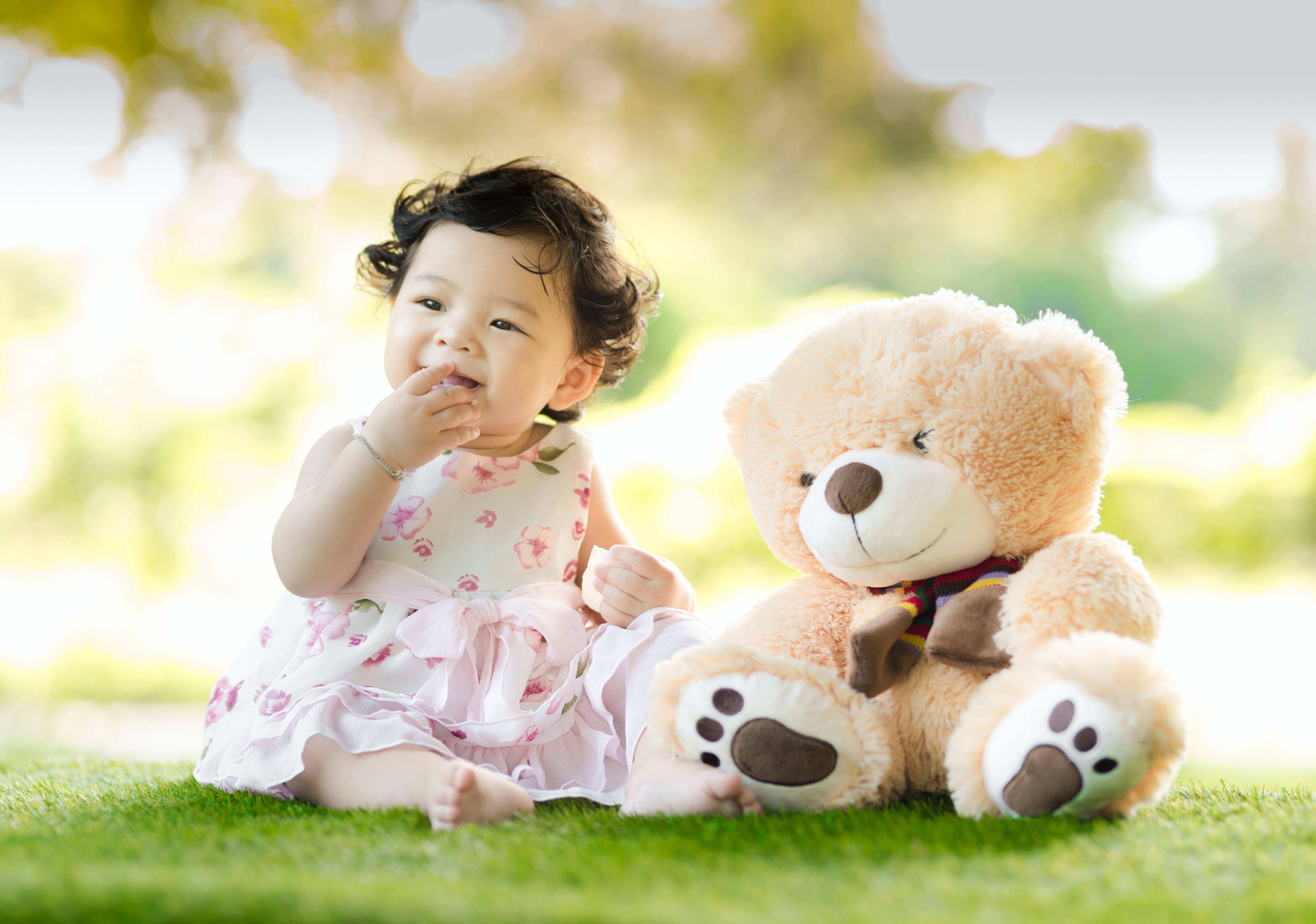 Teddy Bear 桌布 壁紙 — 一個穿著碎花裙的快樂女嬰坐在草地上，旁邊有一隻大泰迪熊，在柔和陽光下。