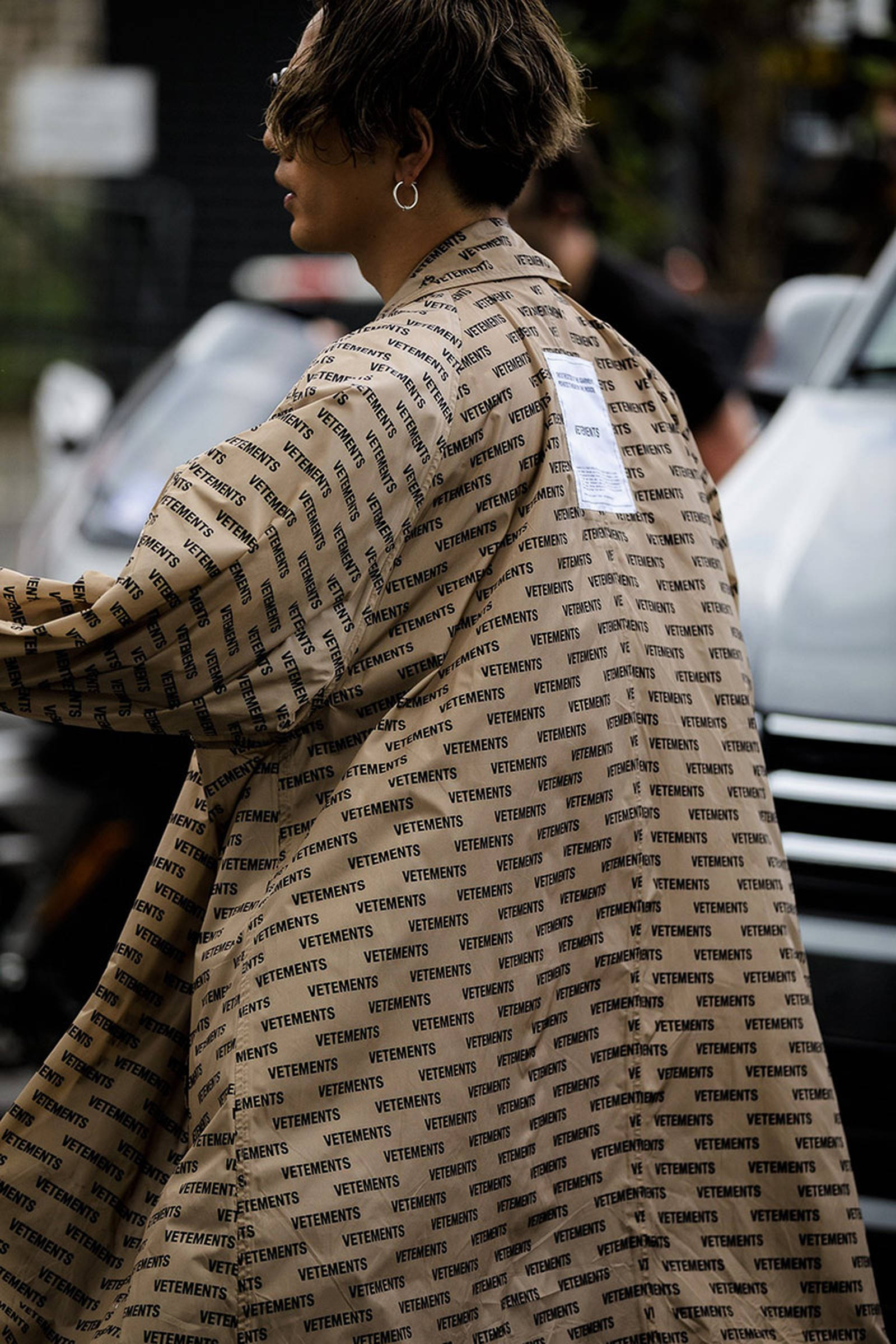 Vetements 桌布 壁紙 — 一個穿著米色VETEMENTS外套、帶有重複文字圖案的人走過模糊的城市街景。