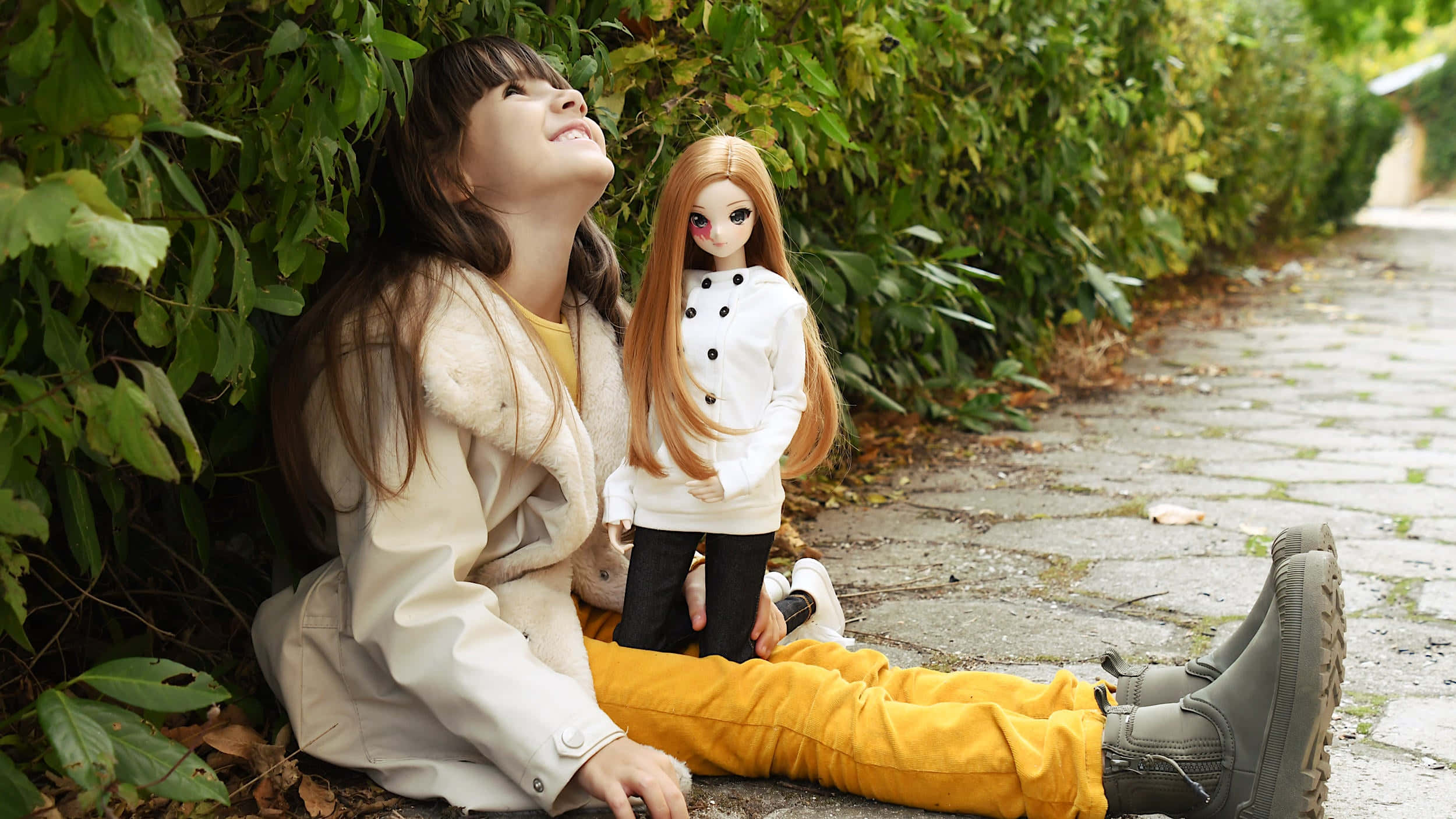 Doll 桌布 壁紙 — 一個穿著米色外套和黃色褲子的小女孩坐在石徑上，旁邊有綠色樹葉，她抱著一個長金髮的洋娃娃，向上微笑。