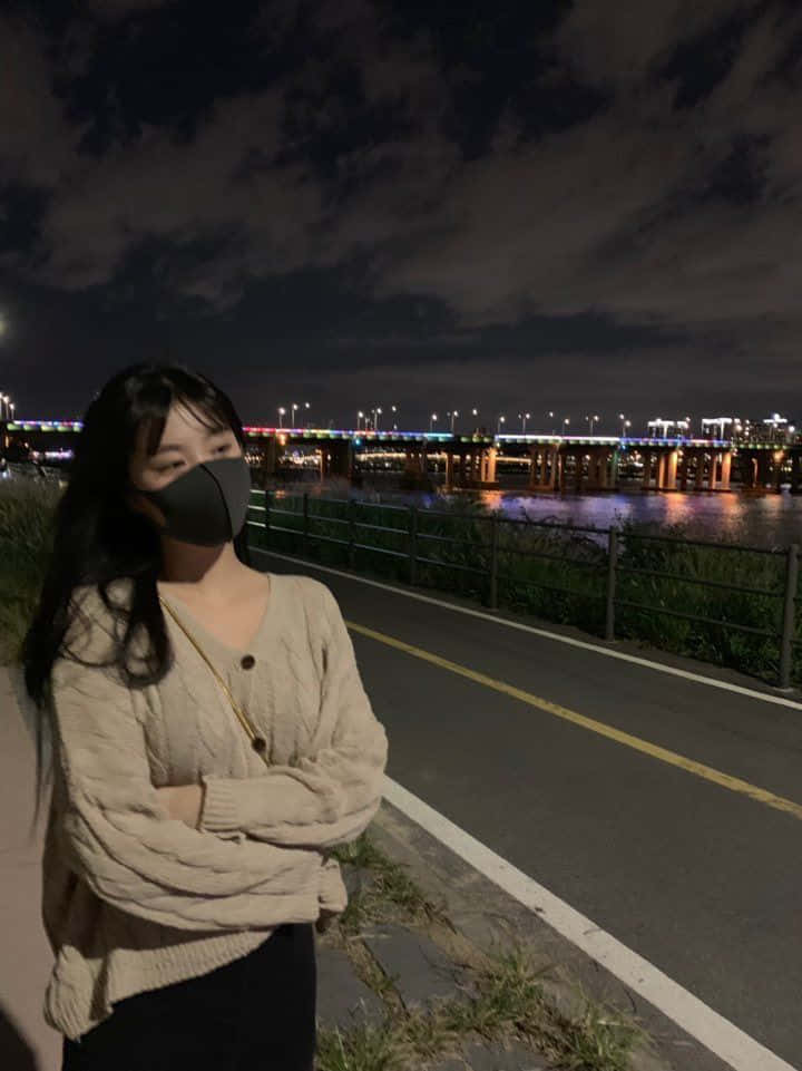 Road 桌布 壁紙 — 一個穿著米色毛衣和口罩的人晚上站在河邊小徑上，背景是一座燈火通明的橋樑。