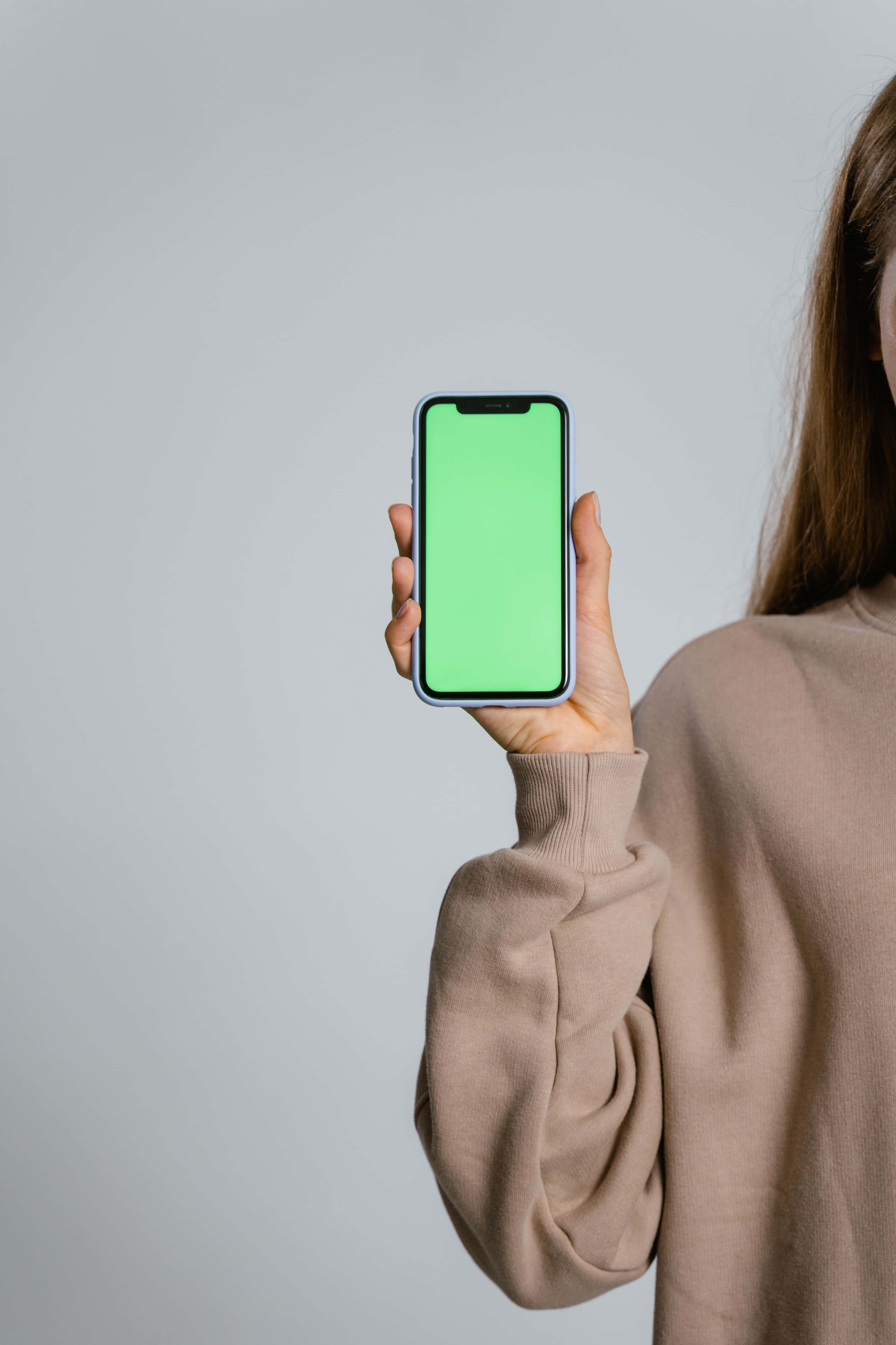 Smartphone 桌布 壁紙 — 一個穿著米色毛衣的人手持一部螢幕亮綠色的智慧型手機，背景為純灰色。