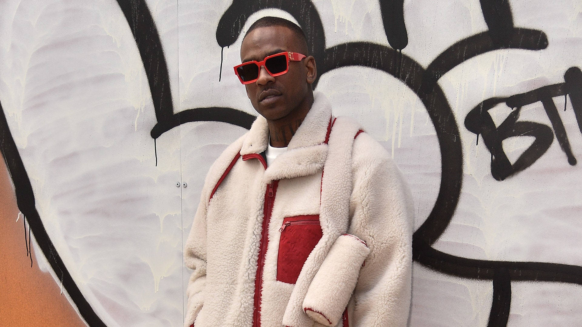 Skepta 桌布 壁紙 — 一個穿著米色羊羔絨外套、帶有紅色點綴和紅色太陽眼鏡的男人，自信地站在佈滿塗鴉的牆前。