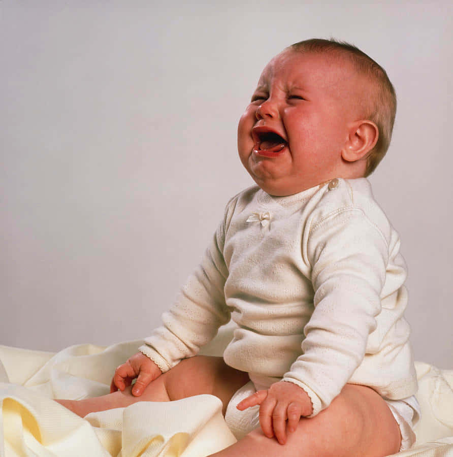 cry 桌布 壁紙 — 一個穿著米色連身衣的嬰兒坐在柔軟的毯子上，緊閉雙眼，張大嘴巴大聲哭泣。