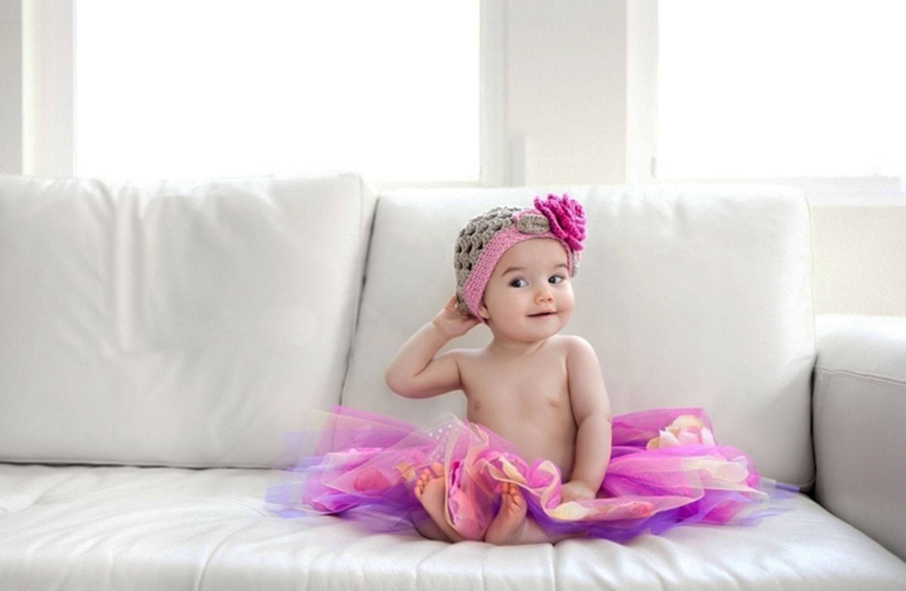 寶寶 桌布 壁紙 — 一個穿著粉紅色和紫色芭蕾舞裙和鉤針頭帶的女嬰坐在白色沙發上，好奇地看著相機。