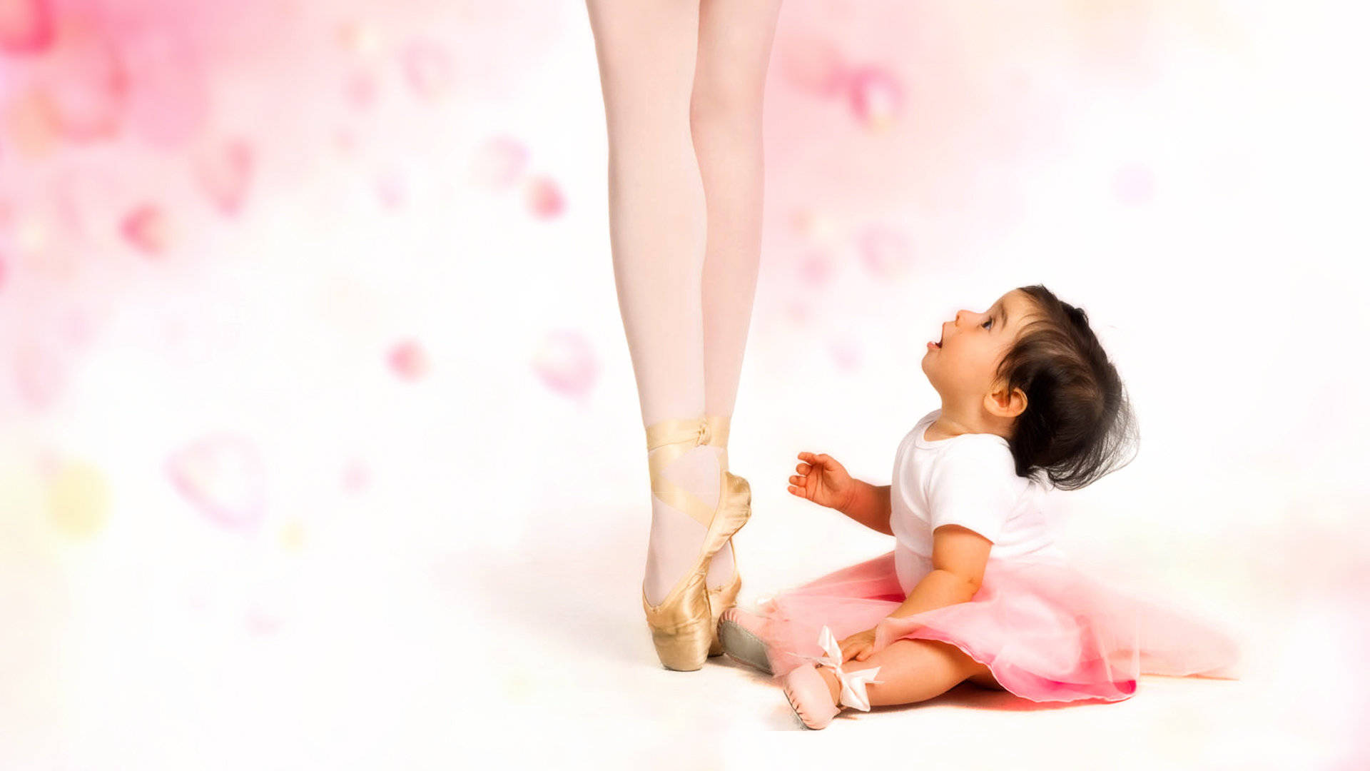 Ballet 桌布 壁紙 — 一個穿著粉紅色芭蕾舞裙的嬰兒，在柔和的粉紅色心形散景背景下，好奇地抬頭看著芭蕾舞者穿著足尖鞋的腳。