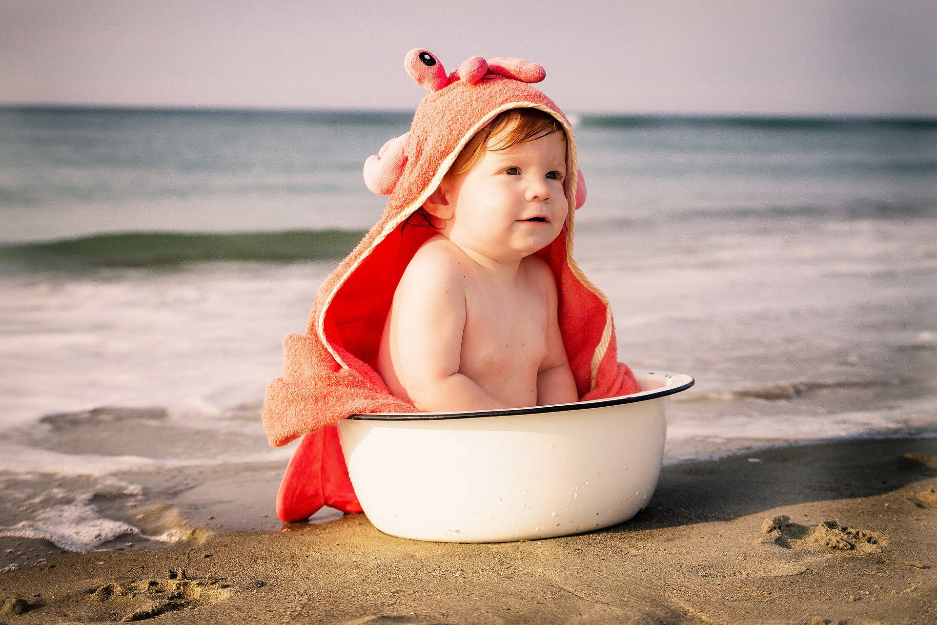 Very Cute Baby 桌布 壁紙 — 一個穿著粉紅色螃蟹主題毛巾的嬰兒坐在沙灘上的白色琺瑯盆中，背景有海浪。