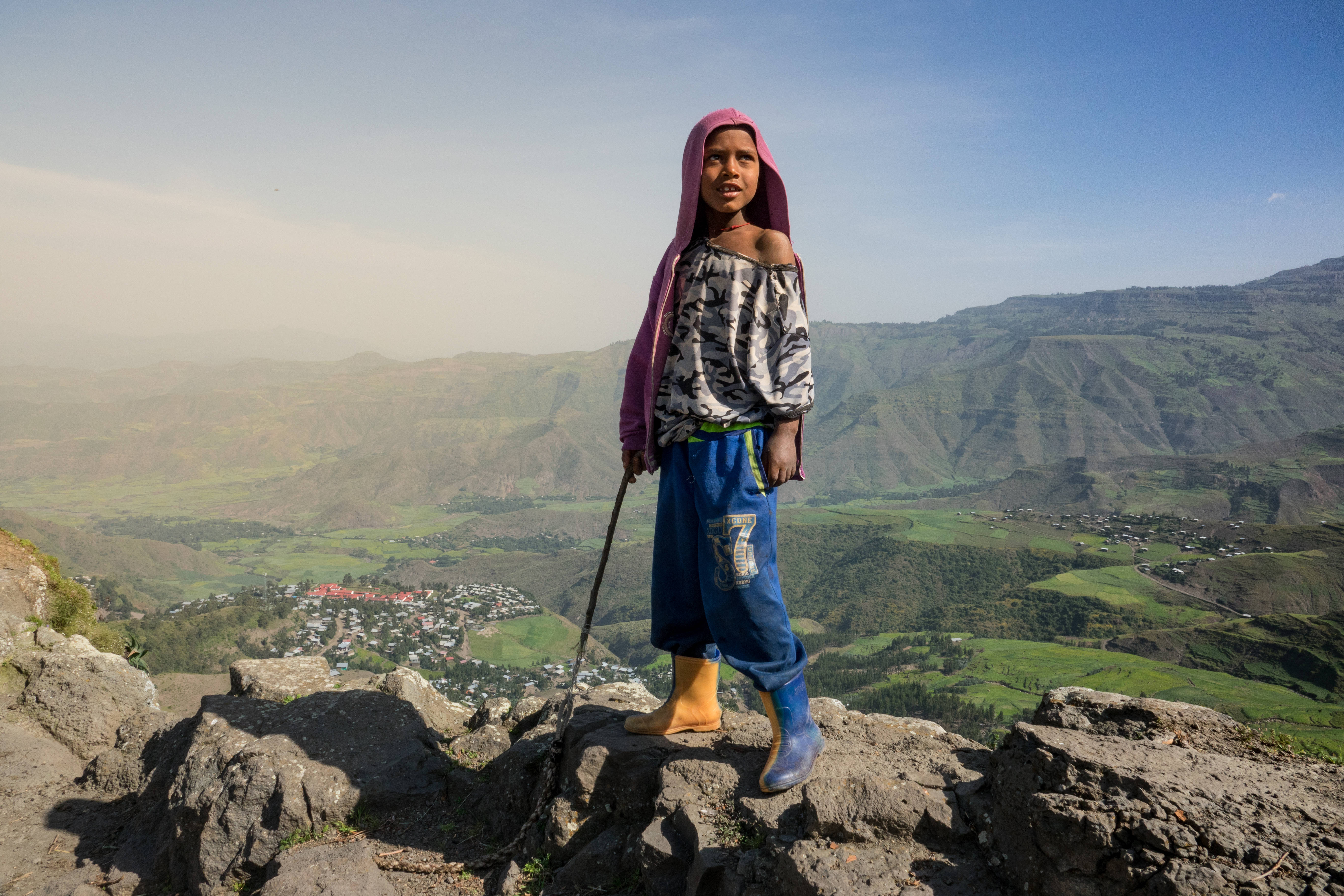 Ethiopia 桌布 壁紙 — 一個穿著粉紅色連帽衫和藍色褲子的小孩站在岩石懸崖上，俯瞰著廣闊的綠色山谷，遠處有山丘，天空清澈湛藍。