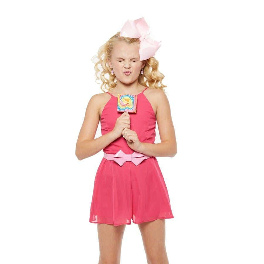 JoJo Siwa 桌布 壁紙 — 一個穿著粉紅色連衣裙、戴著巨大蝴蝶結的小女孩，在白色背景前拿著彩色棒棒糖，做出頑皮的表情。
