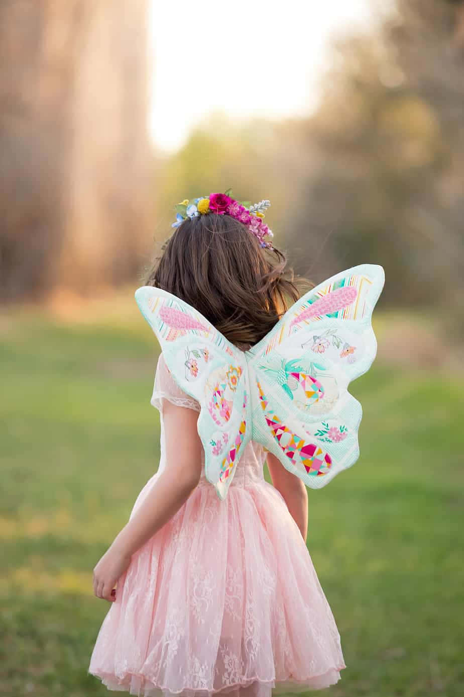 Butterfly Wing Dress 桌布 壁紙 — 一個穿著粉紅色連衣裙和花卉仙子翅膀的小女孩站在陽光明媚的田野中，背景是模糊的森林。