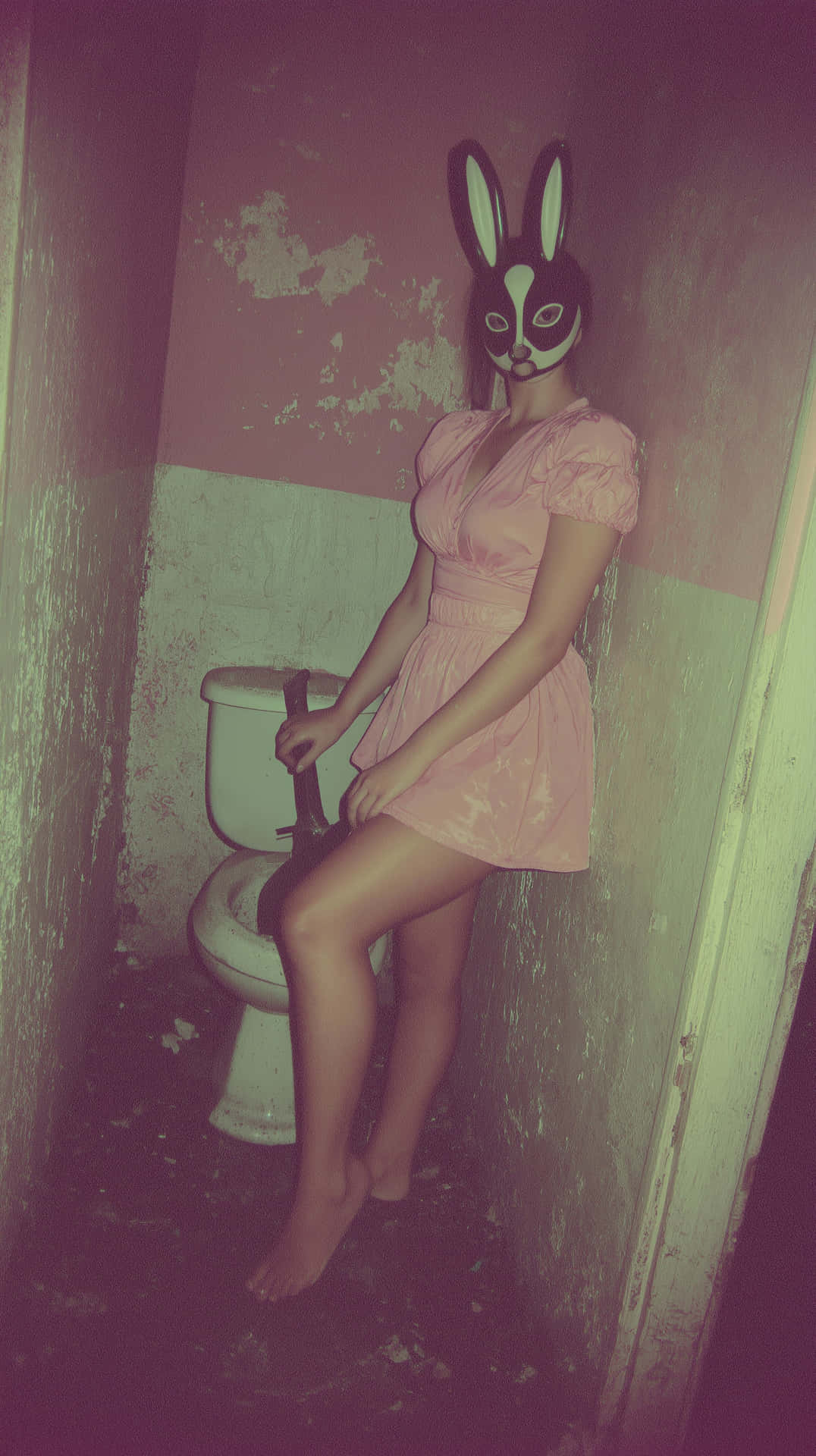 Eerie Rabbit Mask Figure in Derelict Bathroom 壁紙 — 一個穿著粉紅色連衣裙和黑色兔子面具的人赤腳坐在一個牆壁剝落的破舊浴室中的馬桶上。
