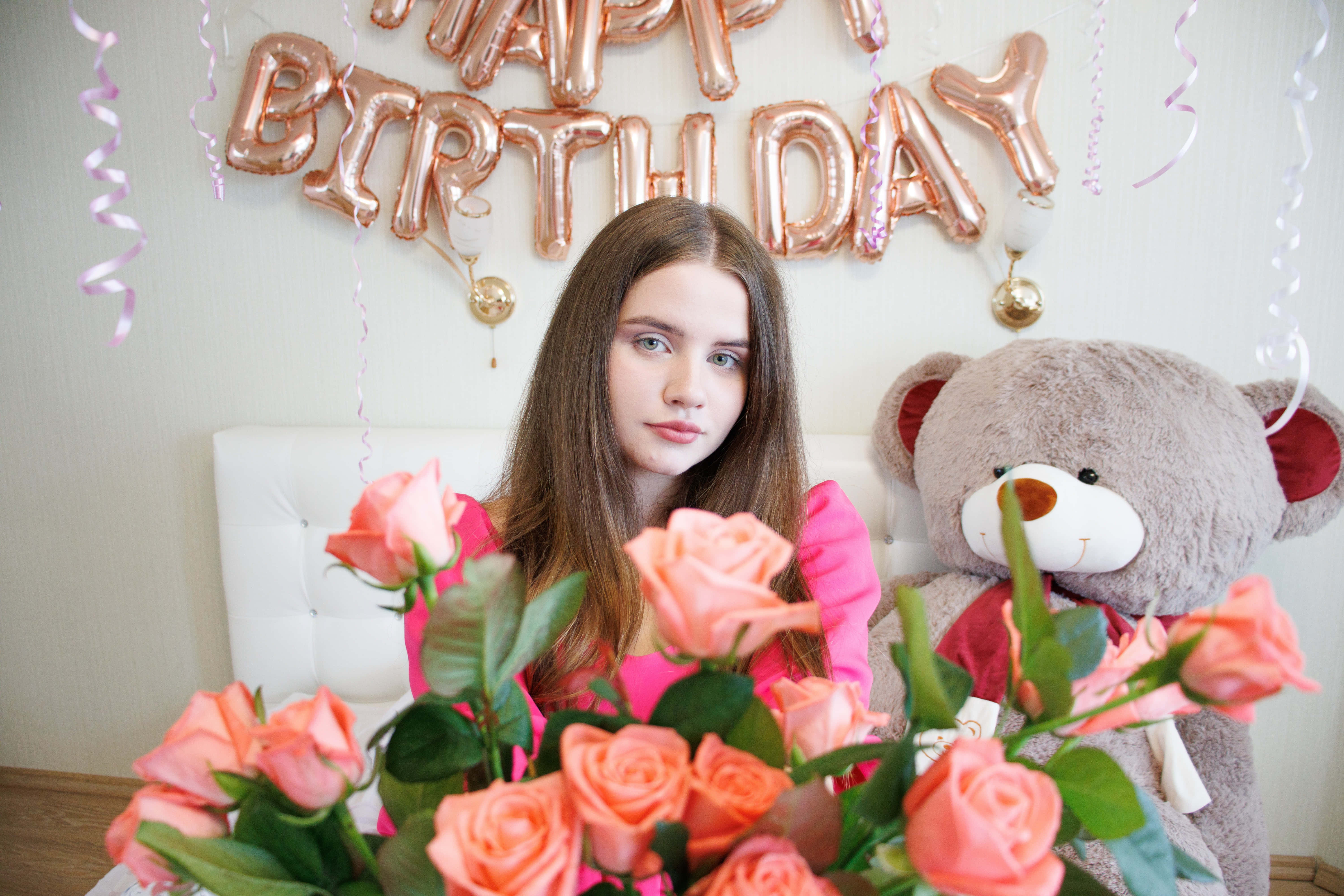 Cute Birthday 桌布 壁紙 — 一個穿著粉紅色連衣裙的小女孩坐在玫瑰金生日氣球下，周圍環繞著蜜桃玫瑰和泰迪熊。
