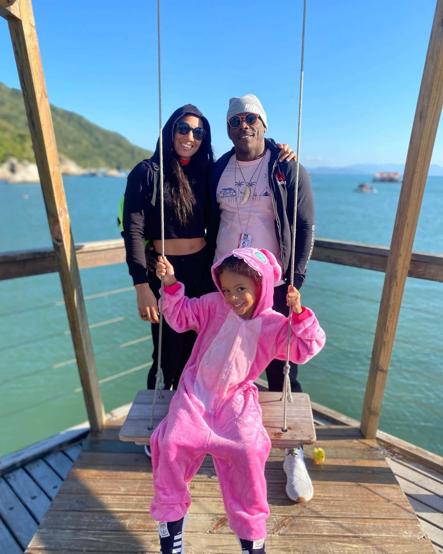 Vacation 桌布 壁紙 — 一個穿著粉紅色連身衣的笑臉小孩與兩個大人坐在陽光明媚的海邊碼頭的木鞦韆上。