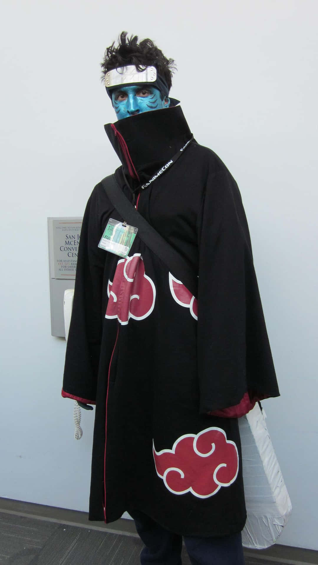 Akatsuki Kisame 桌布 壁紙 — 一個穿著精細曉組織角色扮演、帶有藍色面部彩繪和紅色雲紋斗篷的人，在室內靠著素牆站立。