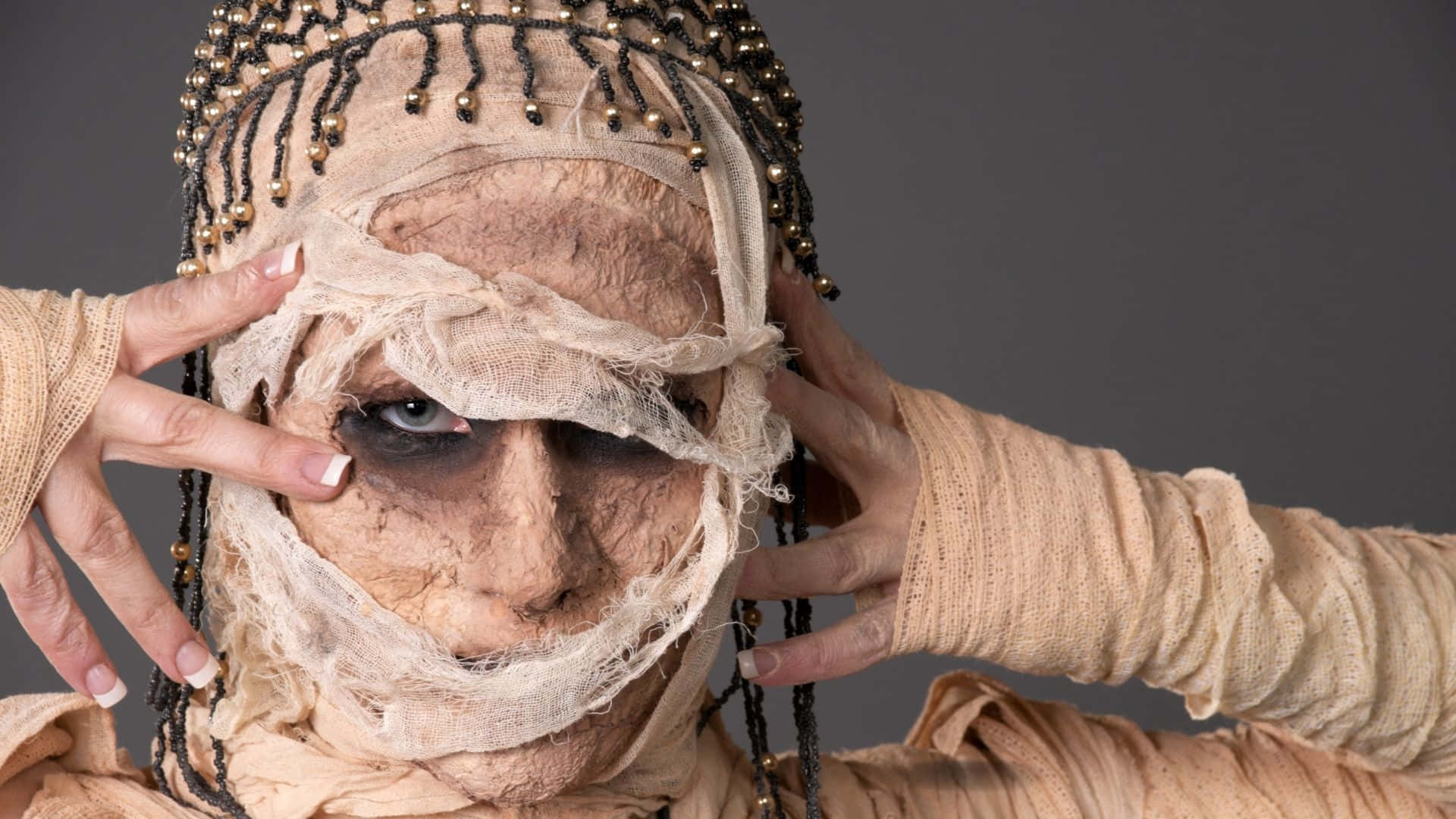 Mummy Costumes 桌布 壁紙 — 一個穿著精細木乃伊服裝的人，臉部和手部纏著繃帶，在灰色背景前掀起布料露出一隻眼睛。