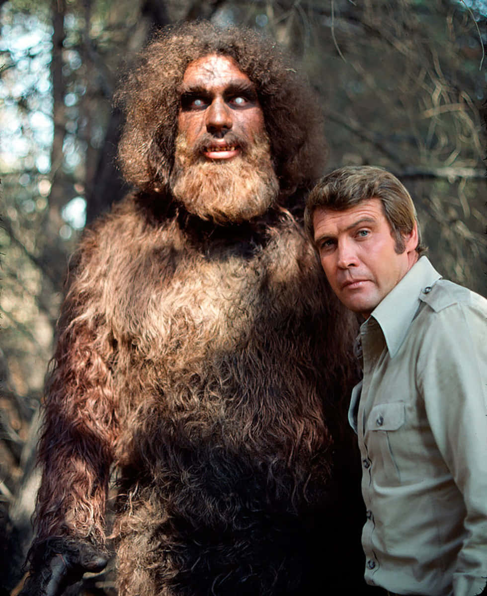 Bigfoot 桌布 壁紙 — 一個穿著精細毛茸茸大腳怪服裝的男人，在森林環境中站在一名人類演員旁邊。