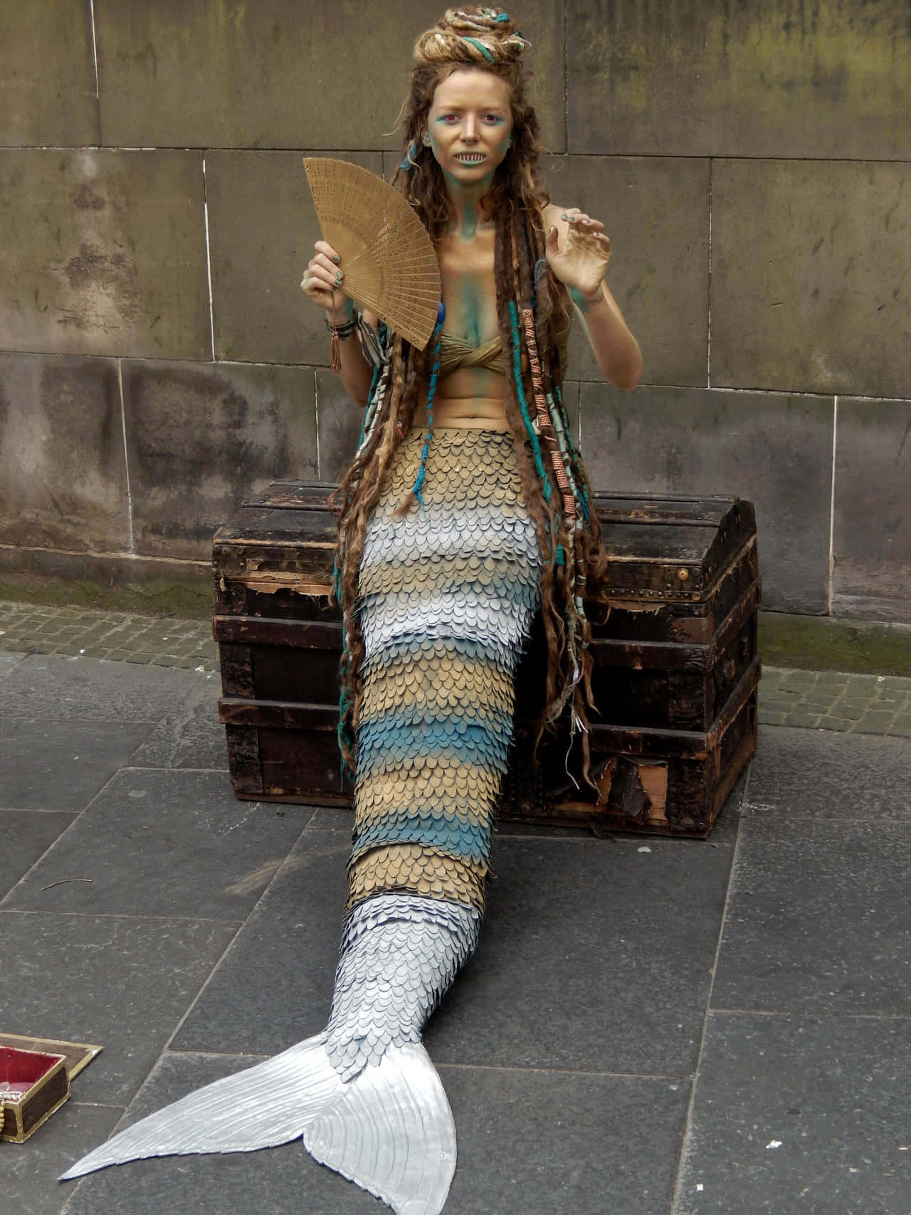 Real Mermaid 桌布 壁紙 — 一個穿著精細美人魚服裝、帶有鱗片圖案尾巴和編織配飾的人，坐在靠石牆堆疊的木箱上。