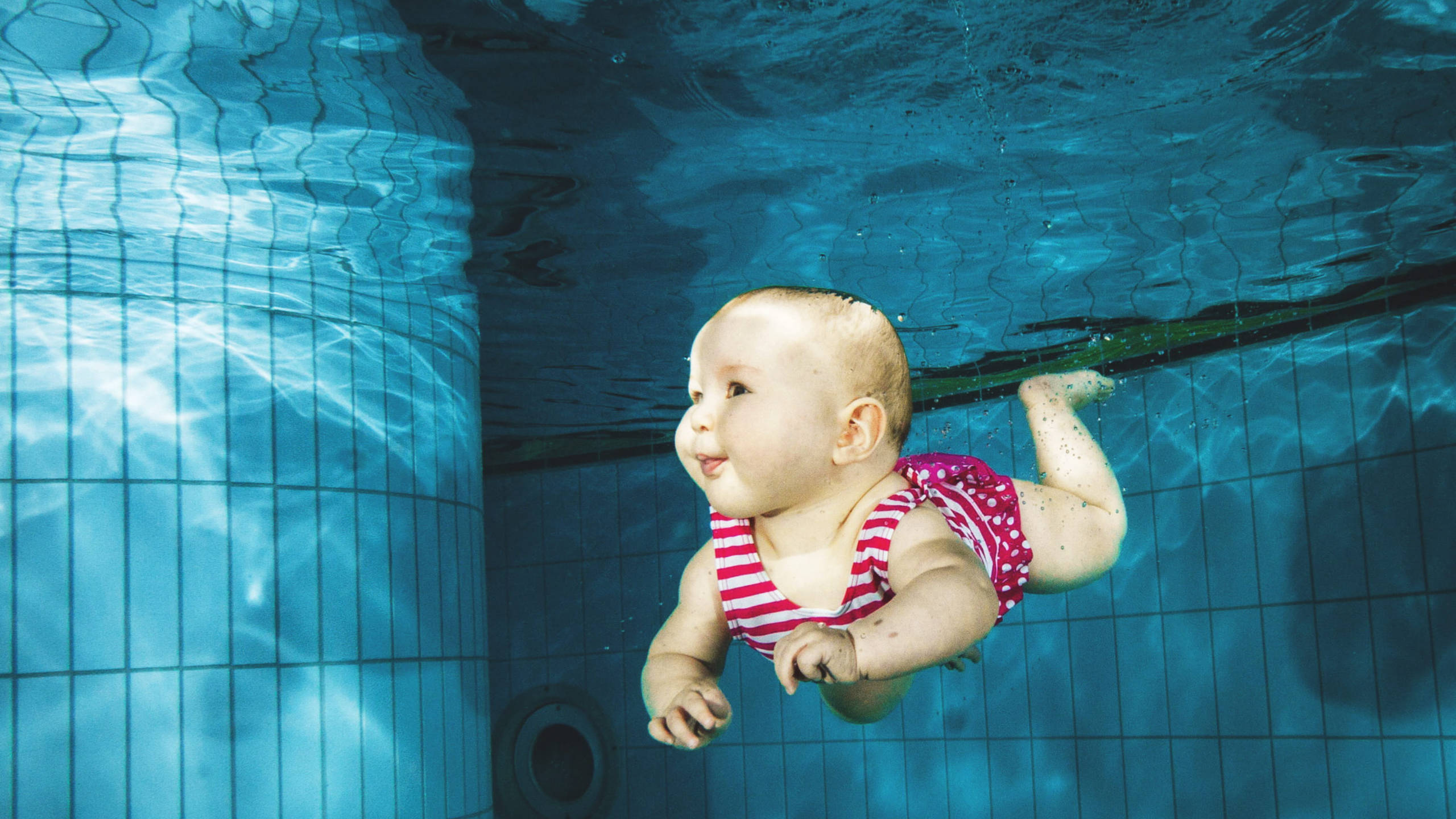 Swimming 桌布 壁紙 — 一個穿著紅白條紋泳衣的嬰兒在藍色瓷磚泳池的水底漂浮，陽光穿過水面照射下來。