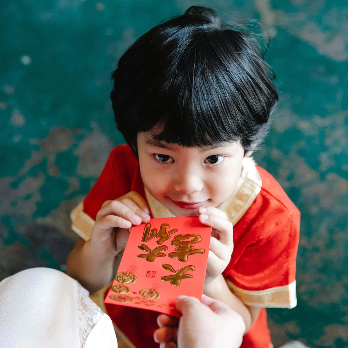 中文 桌布 壁紙 — 一個穿著紅色傳統服裝的小孩拿著一個帶有金色字樣的紅色紅包，抬頭露出溫柔的微笑。